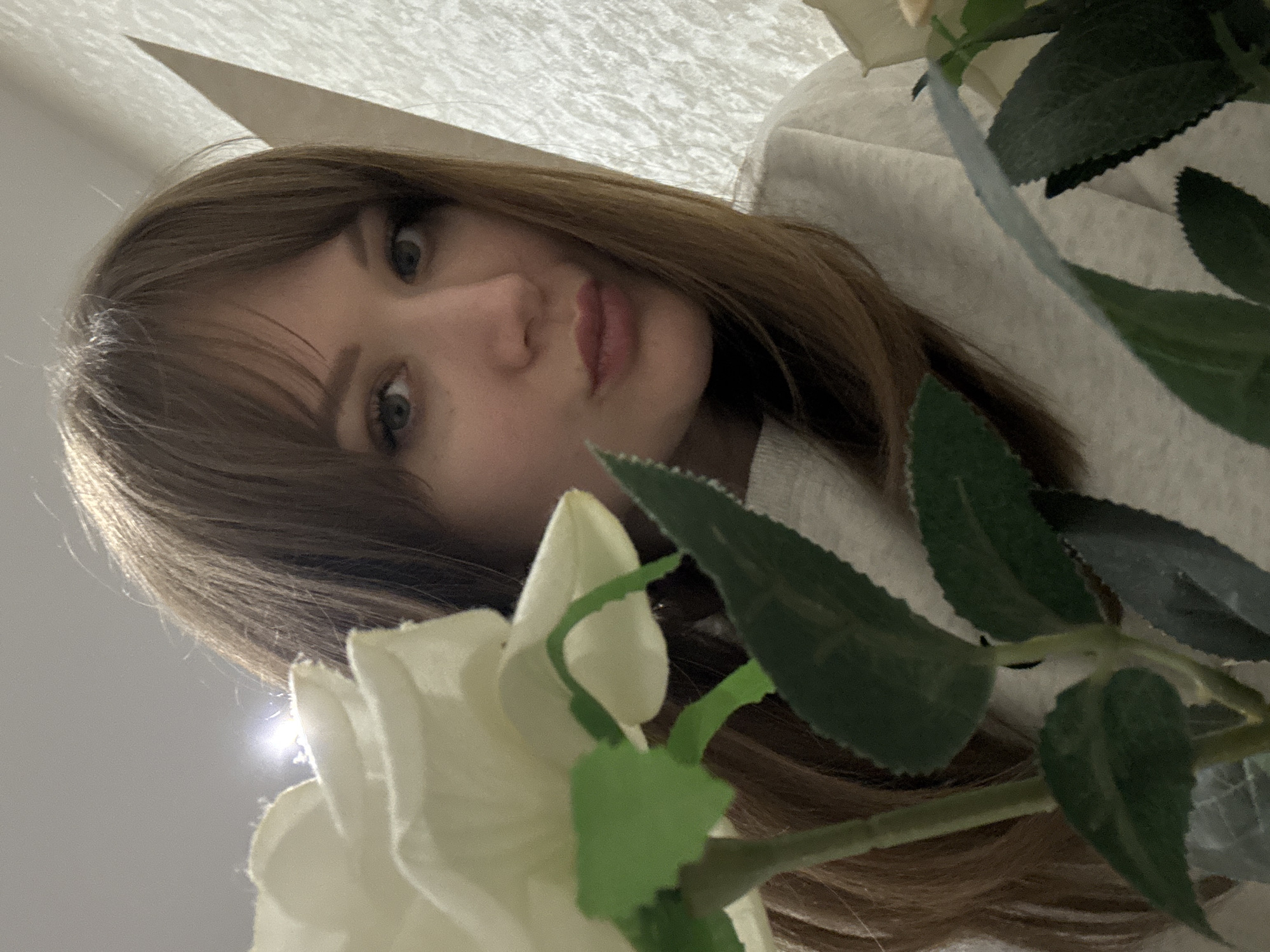 A young woman with brown hair and fair skin is posing close to the camera with a slightly pursed expression. She is wearing a light-colored garment. In the foreground, artificial white flowers and green leaves are partially obscuring the view. The background appears to be a textured white surface, possibly a wall or fabric. There is no evidence to suggest the location is Saratov, Russia.