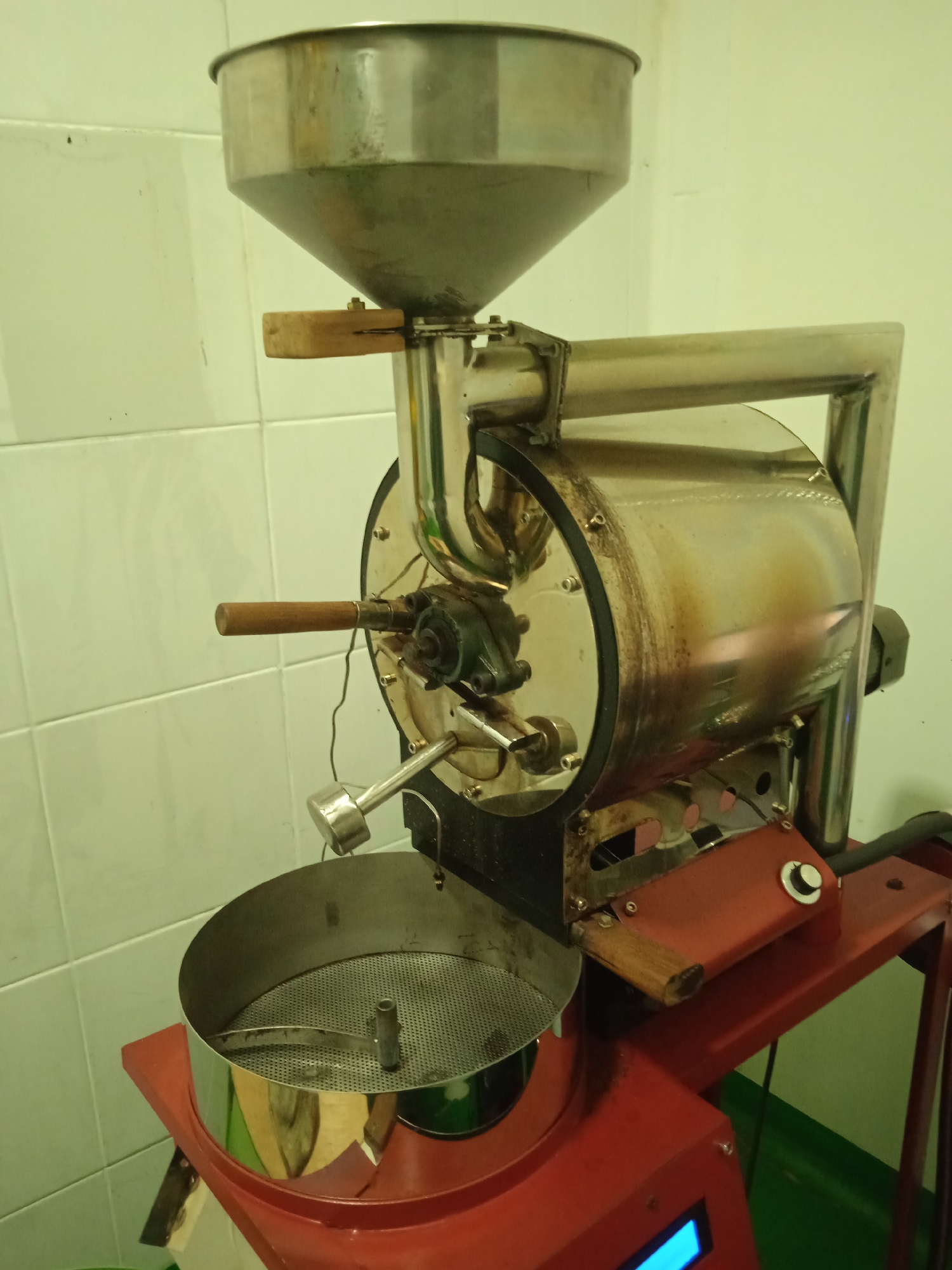 A coffee roasting machine is shown in a room with light green tiled walls. The machine is primarily made of stainless steel and painted red. At the top of the machine is a large, funnel-shaped hopper. Below the hopper, a rotating drum is visible, where the coffee beans are roasted. A wooden handle protrudes from the side of the drum. Beneath the drum is a cooling tray with a perforated surface. The machine is situated on a red metal stand, and a small blue digital display is visible on the front of the stand. The overall impression is of a functional piece of equipment in a commercial or specialized setting. The location is identified as Cileunyi, Indonesia.