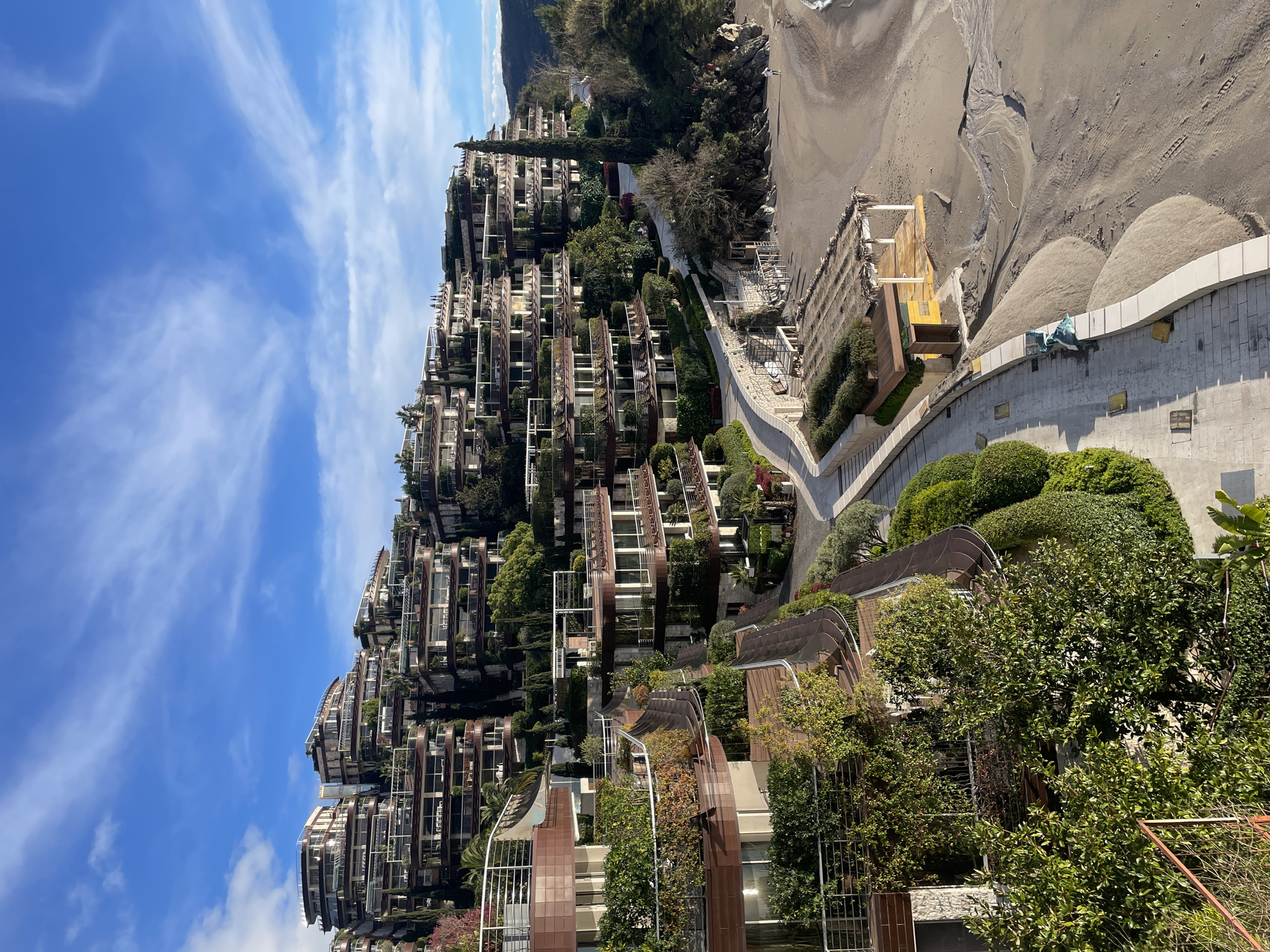 The image depicts a panoramic view of a terraced architectural complex descending a lush hillside towards a sandy beach and the Adriatic Sea, specifically located in Budva, Montenegro.
The upper and central portions of the frame are dominated by numerous multi-story buildings, characterized by their curved, staggered design with extensive balconies and integrated planters. These structures feature facades with warm brown tones, contrasted by white railings, and are heavily adorned with green vegetation, including various shrubs, small trees, and climbing plants. Tall, slender coniferous trees, resembling cypress, punctuate the landscape between the buildings and on the right side.
Below the main architectural complex, a paved stone walkway curves along the coastline, bordered by low, light-colored walls and manicured green hedges. Adjacent to this walkway lies a light-brown sandy beach. The beach exhibits areas of disturbed sand, including small ridges and channels indicative of water flow, and faint tracks.
On the beach, near the walkway, there is a small, open-sided structure with a thatched roof supported by light-colored poles. Attached to this structure is a smaller, enclosed yellow shed. Further along the beach, partially obscured, is what appears to be a disused or partially disassembled wooden platform. Near the retaining wall separating the walkway from the sand, several green-colored bags are visible.
On the far right of the beach, a single individual, wearing light-colored clothing, is seen walking near the waterline. The water of the sea is visible on the far right, with a gentle wave line meeting the shore. Beyond the immediate scene, natural hills covered in sparse vegetation rise in the background.
The sky is bright blue with scattered, wispy white clouds. The scene is illuminated by clear daylight, with shadows suggesting the sun is positioned to the left and slightly behind the vantage point. No overt actions or interactions between individuals are depicted, beyond the single person walking.