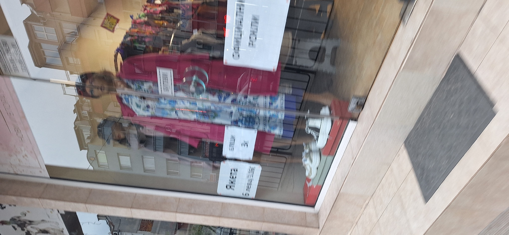 The image shows a shop window in Blagoevgrad, Bulgaria. A red and blue patterned jacket is displayed in the window, alongside a sign written in Bulgarian. The reflection in the glass reveals buildings across the street and a person taking the picture. The sidewalk has gray carpet.