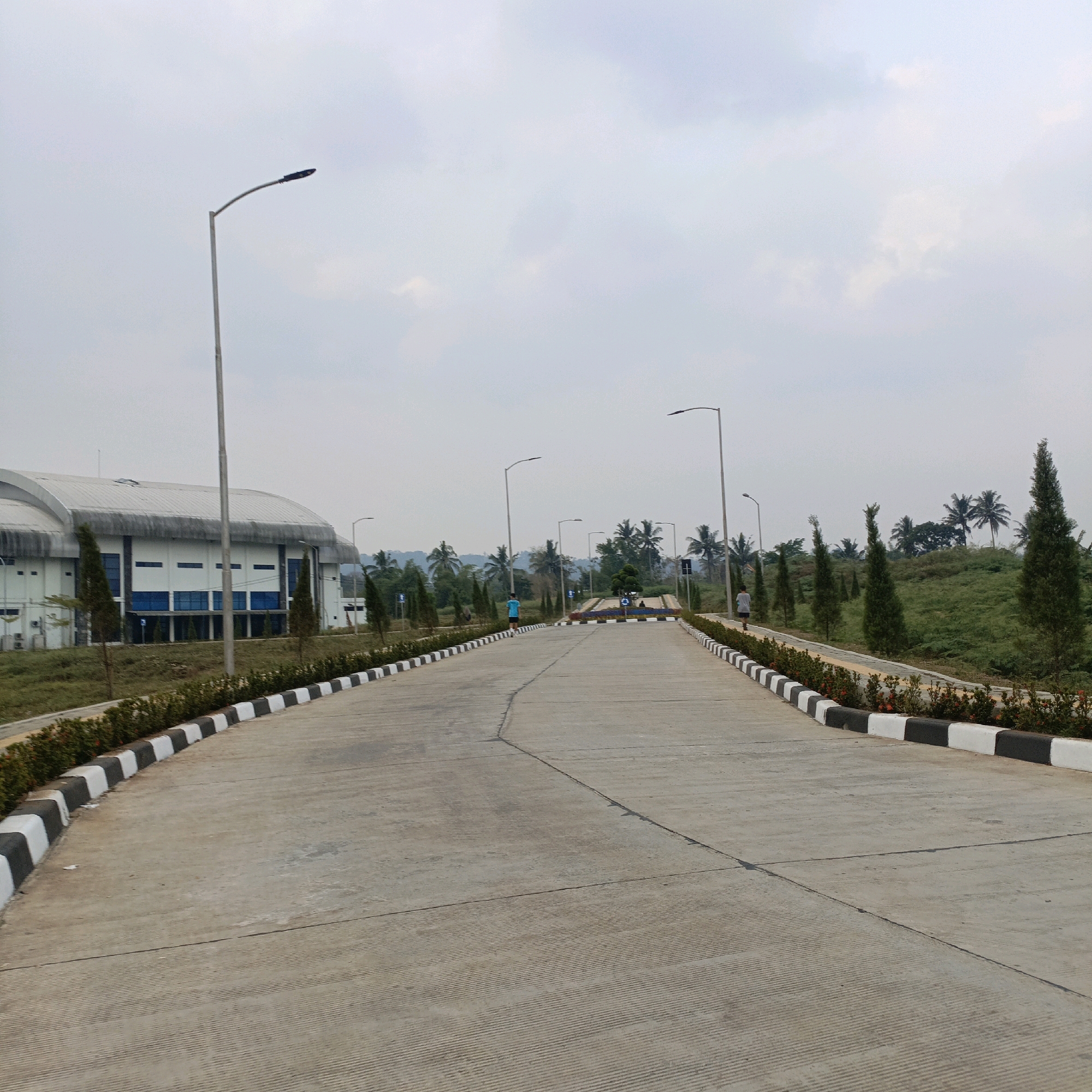 The image shows an exterior view of a modern industrial or institutional complex under a cloudy sky. A wide, paved road with distinct black and white striped curbs curves through the scene. On the left, a large, white building with a curved, silver roof and blue-tinted windows is visible. Several tall, slender cypress trees are planted along the road, interspersed with green foliage and flowering bushes in the foreground. In the background, palm trees and lush green vegetation are visible, suggesting a tropical or subtropical climate. Two individuals are seen walking or jogging on the road, one in a blue t-shirt and the other in a grey t-shirt, appearing to be engaged in recreational activity. Streetlights are positioned along the road. The overall impression is one of an organized and well-maintained outdoor space. Based on the provided context, this scene is located in the city of Salatiga, Indonesia.