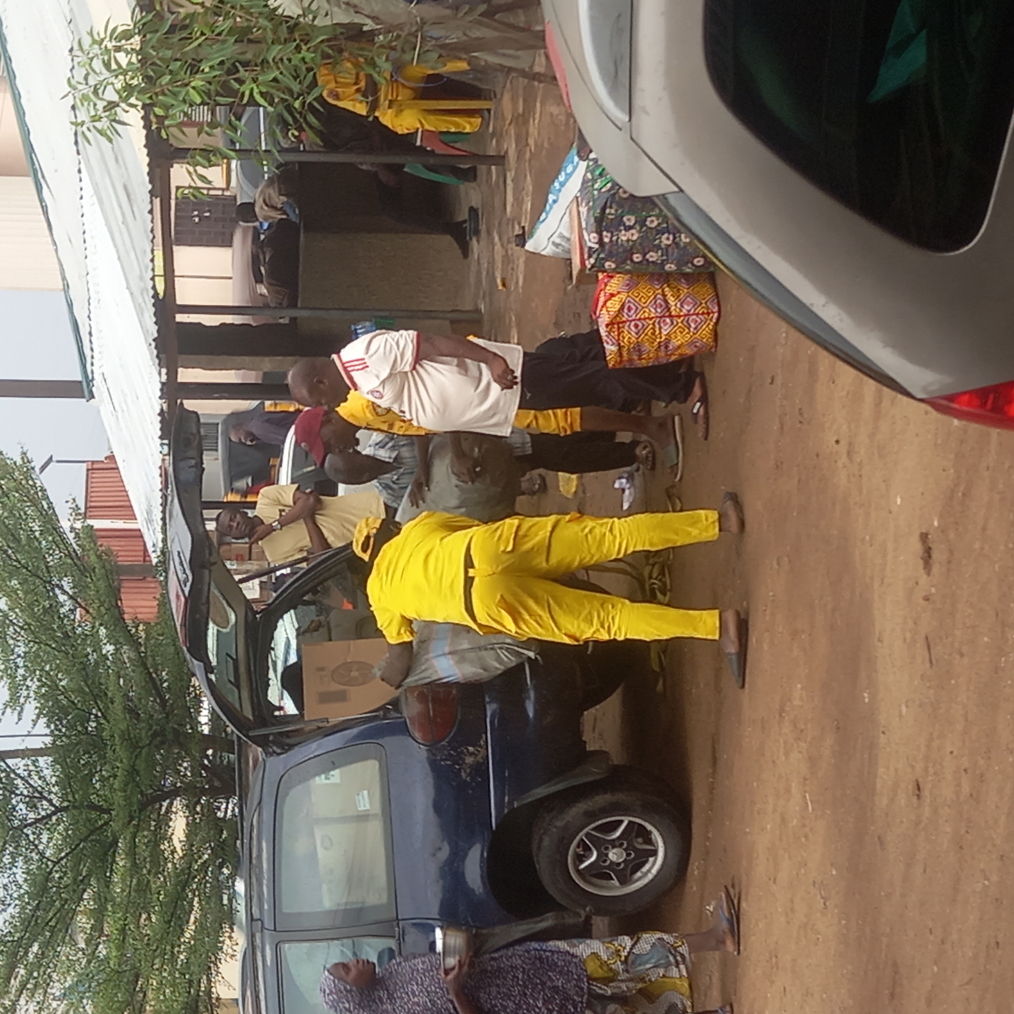 The image shows several people in what appears to be a loading or unloading process near a dark blue car, likely in Sardauna, Nigeria. The car's trunk is open, and items are being moved into or out of it. Some individuals are dressed in yellow outfits. The environment consists of a dirt ground area near buildings with corrugated roofing. A silver vehicle is parked partially obstructing the view of the scene. The scene has a somewhat disorganized feel, with people standing around and engaged in various actions.