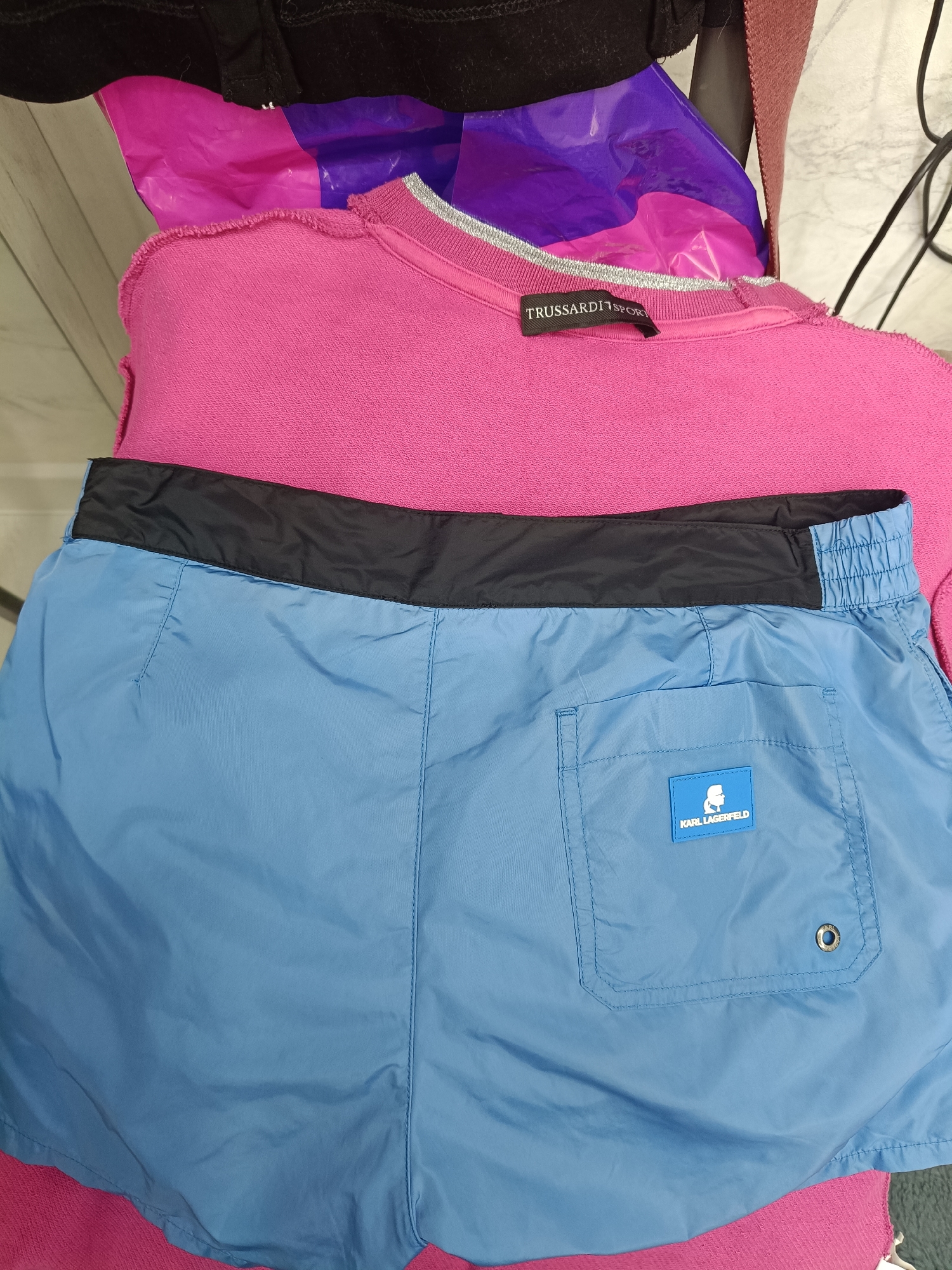 A close-up, top-down view shows two articles of clothing laid out on what appears to be a flat surface. The upper garment is a bright pink sweatshirt or sweater with a raw hem. A black tag with "TRUSSARDI SPORT" printed in white is visible on the neckline. Beneath the pink garment are light blue shorts. The shorts have a black waistband and a visible pocket on the right side, adorned with a blue label featuring the Karl Lagerfeld logo and text. The background is blurred and appears to be an indoor setting with hints of a marble surface and some dark objects. The image is well-lit, and the colors are vibrant. The location of Sochi, Russia, is not evident from the image.