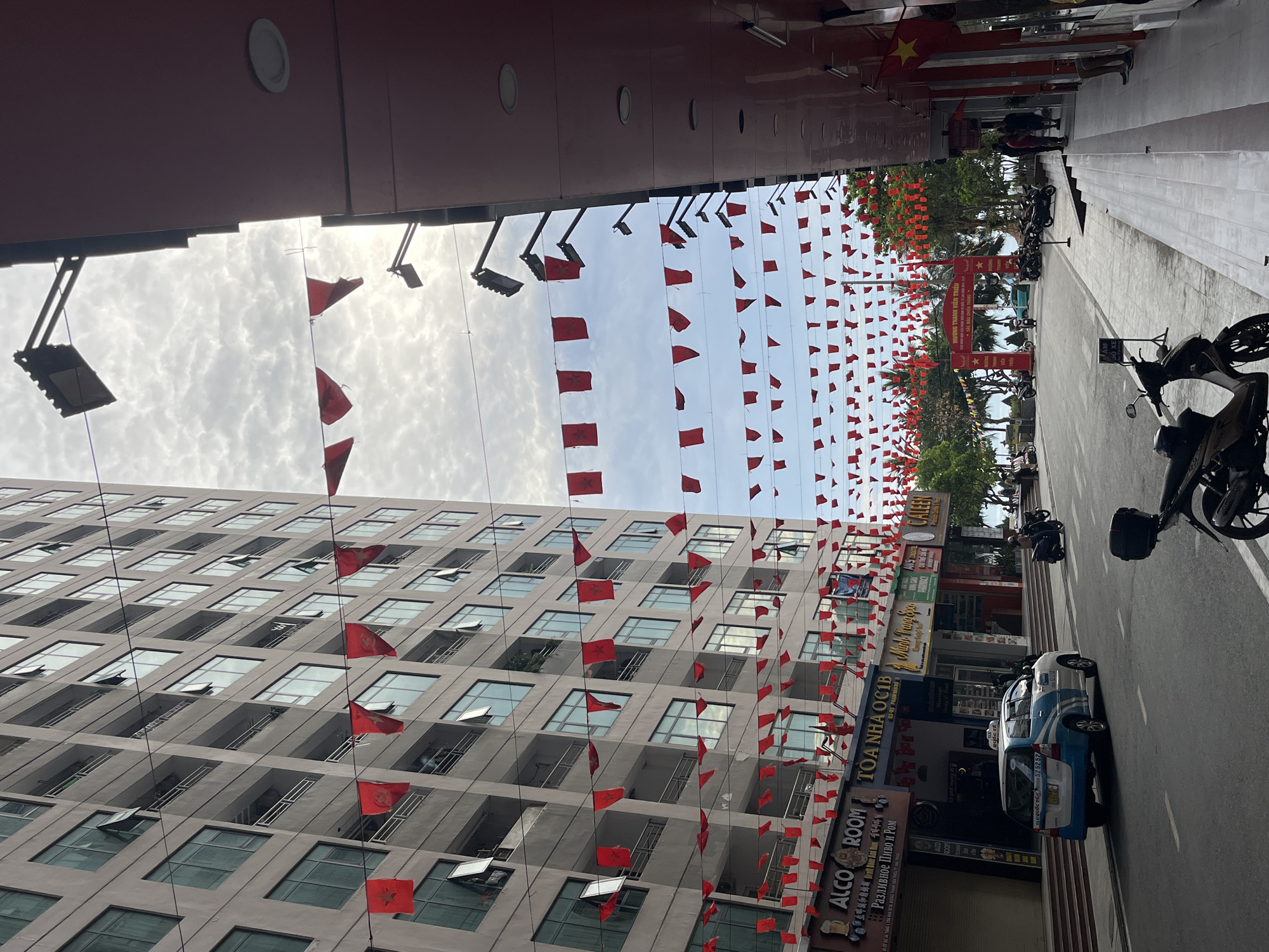 The image depicts a street in Bac Nha Trang Ward, Vietnam, characterized by tall buildings adorned with numerous red Vietnamese flags strung overhead. A multi-story building on the left exhibits a facade with numerous windows. The street itself has parked motorcycles, one prominently in the foreground, and a blue and white taxi. Storefronts are visible on the right side of the street with signs in Vietnamese and other languages, with "ALCO ROOM" and "TOA NHA OC1B" visible. Pedestrians are present near the end of the street. The sky above is partly cloudy.