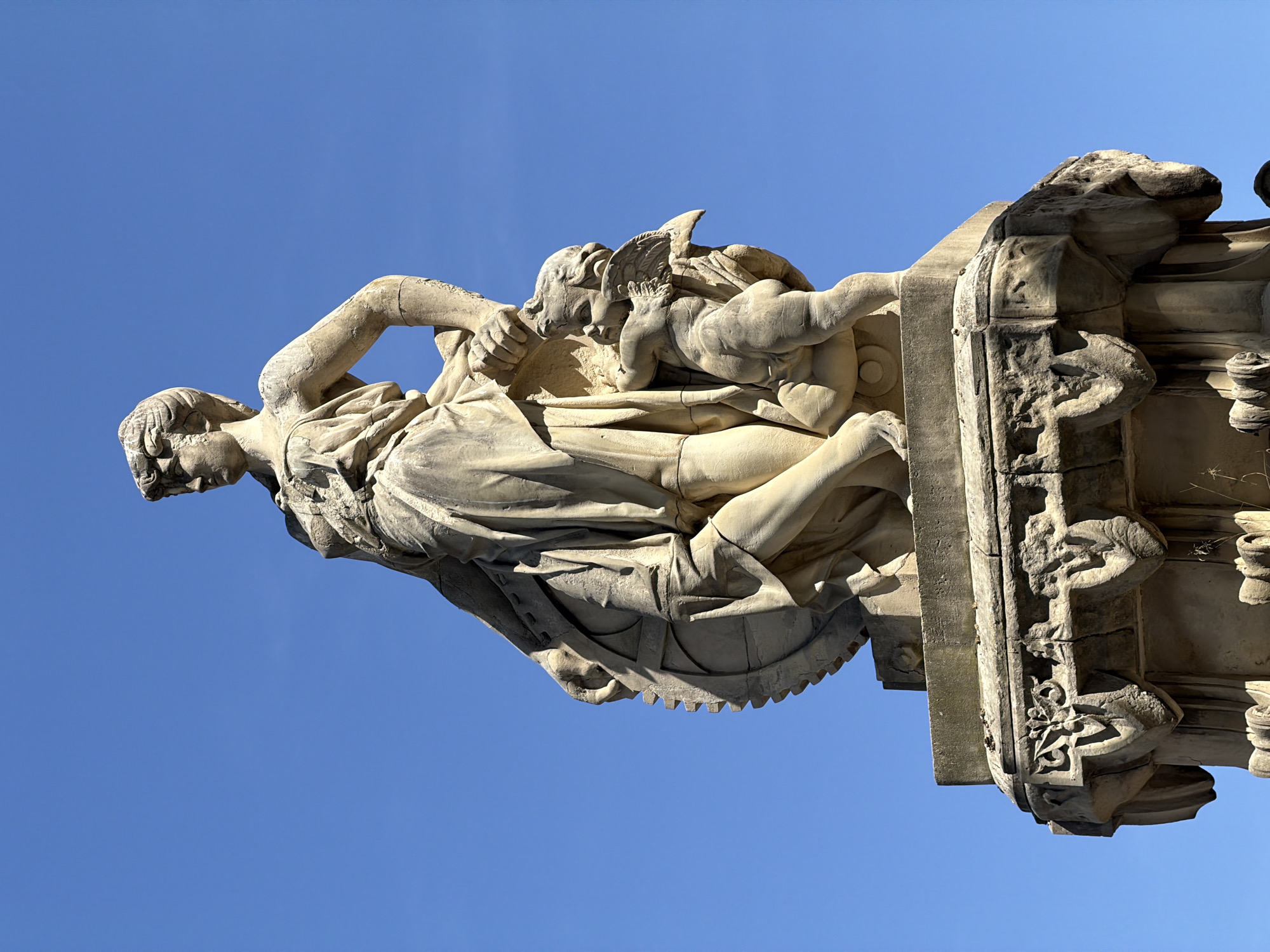 The image shows a stone statue of a female figure and a cherub against a clear blue sky. The statue is mounted on an ornate stone pedestal. The female figure is depicted wearing a draped garment, and her hair is styled in an updo. She is looking to her left, with her left arm bent and her hand resting on a large circular object with a toothed edge. A cherub with outstretched wings stands to her right, looking up at her. The pedestal features decorative carvings, including floral motifs and grotesque faces. The statue appears to be made of weathered stone and is set against a bright, cloudless sky. This statue is located in the Parc de la Ciutadella in Barcelona, Spain.