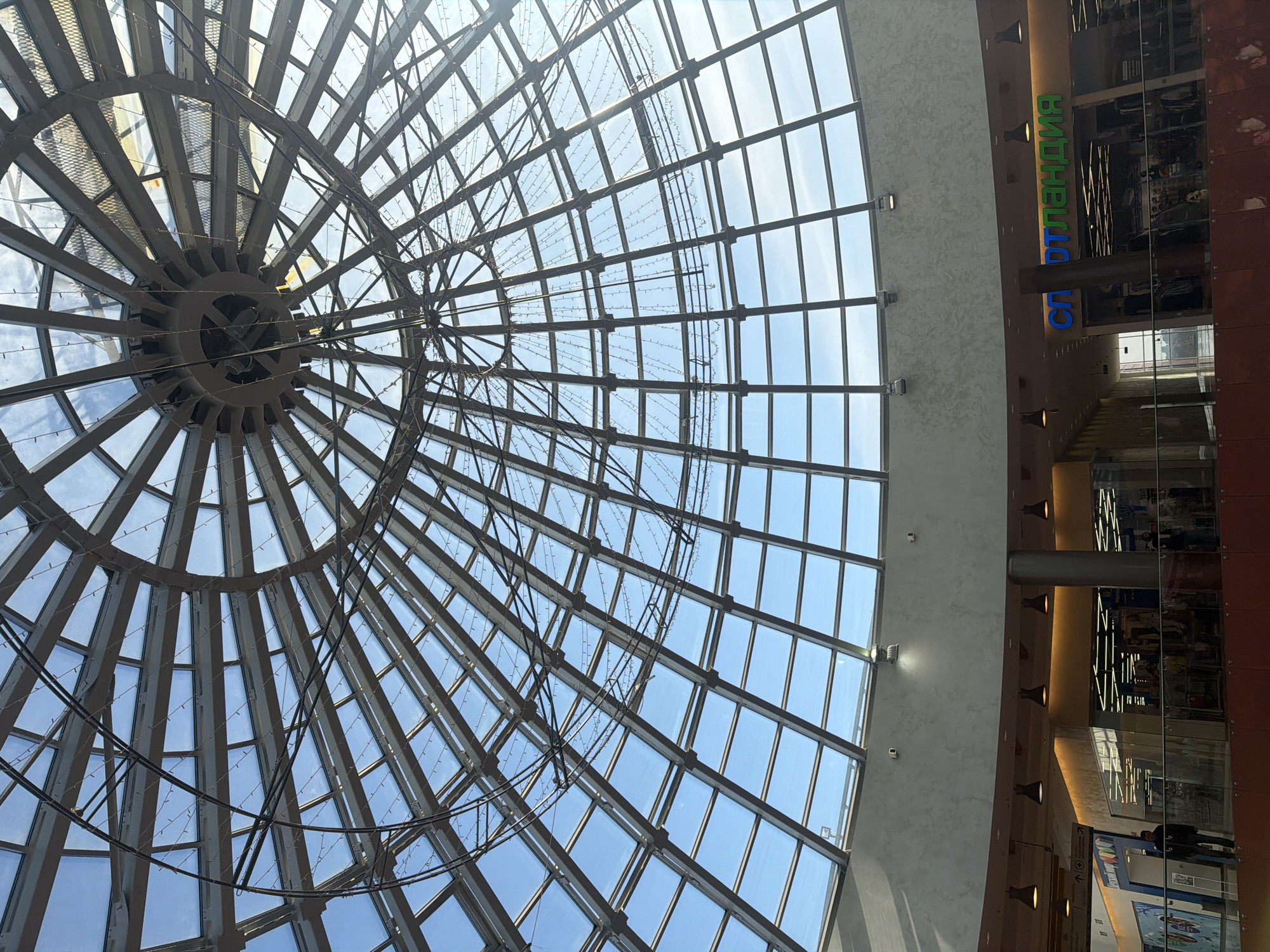 The image captures the interior atrium of the Semya (Семья) Shopping and Entertainment Complex, located in Perm, Russia. The primary focal point is a large, glass-paneled domed skylight forming the building's roof, structured with a radial and concentric framework of dark metal beams. Numerous rectangular and trapezoidal glass panes allow natural light from a clear blue sky to illuminate the space. Strung across the internal metal framework of the dome are multiple strands of small, white decorative lights.
Below the skylight, a curved, light-colored architectural element, possibly an upper floor balcony or ceiling, is visible, equipped with recessed lighting fixtures and two mounted security cameras. A lower retail level is discernible, featuring a railing composed of a reddish-brown opaque lower section and a transparent upper panel. On this level, storefronts are visible. To the right, a store named "СПОРТЛАНДИЯ" (SPORTLANDIA) is clearly marked with large blue and green Cyrillic letters, showcasing merchandise through its glass facade. To the left, a partial sign for "ДЕТСКИЙ МИР" (CHILDREN'S WORLD) is visible, accompanied by an directional arrow. Approximately five individuals are present on the lower level, casually walking or standing, with no specific interactions or actions discernible. Artificial lighting supplements the natural light, emanating from store interiors and suspended pendant fixtures beneath the curved architectural element.