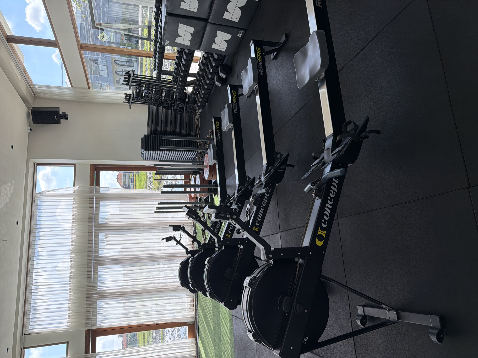 The image captures the interior of a brightly lit fitness gym or studio, oriented with its long axis along the windowed wall. No people are present, indicating a moment of quiet between activities.
The primary subjects are a line of at least five black Concept2 RowErg rowing machines, prominently displayed with their "Concept2" and "RowERG" branding in yellow text. These machines are arranged side-by-side on dark rubber gym flooring, facing large windows that offer an expansive view of the outdoors.
To the left of the rowing machines, a section of the gym is dedicated to weight training. This area features a rack of dumbbells of various sizes, stacked weight plates, and several black plyometric boxes. The plyometric boxes bear a white "S" logo, and some are marked with "20KG."
The gym is well-lit by natural daylight streaming through tall, wide windows that span the length of the room. Some windows are covered with translucent white horizontal blinds or sheer curtains, while others offer a clear view. The window frames appear to be made of wood. Through the clear window sections, a bright blue sky with scattered white clouds is visible, suggesting a clear daytime, likely morning or afternoon. The outdoor scene reveals lush green vegetation, possibly rice paddies, consistent with the North Kuta, Indonesia location, along with some distant buildings. A yellow road sign with an indistinguishable black symbol is also faintly visible outside.
On one of the white walls, a black speaker is mounted, with a small CCTV camera positioned nearby. The overall aesthetic suggests a modern and well-equipped facility designed to incorporate the natural surroundings.