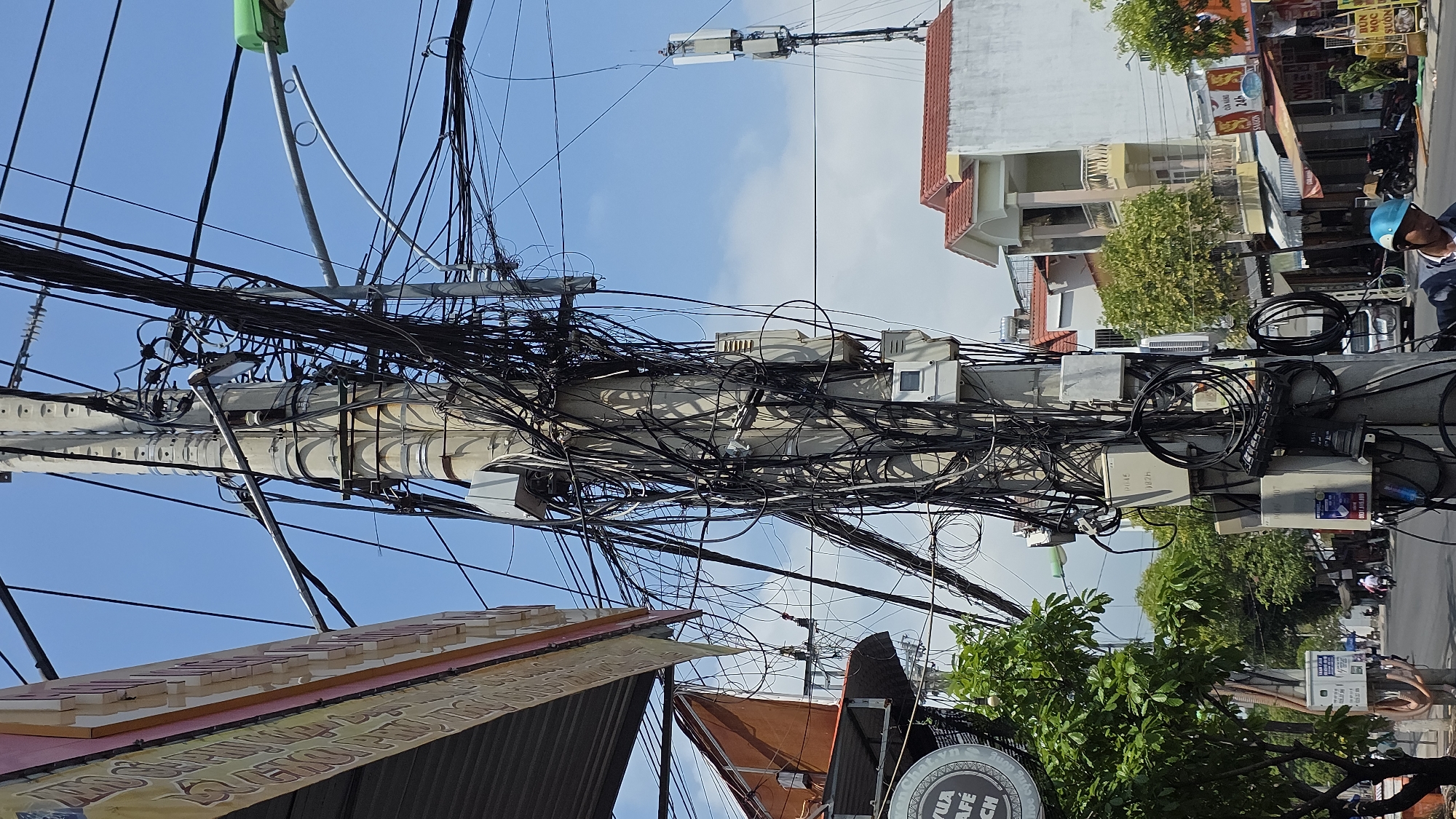 The scene captures the chaotic arrangement of utility wires and electrical boxes densely attached to a concrete utility pole in a street in Nha Trang, Vietnam. The perspective is angled upwards, showcasing the cluttered web of black wires against a partly cloudy blue sky. Adjacent to the pole are buildings with varied architecture, including a three-story structure with a red-tiled roof. Signage and canopies from local businesses are visible. Pedestrians are present, including a person wearing a blue helmet riding a motorbike. The overall impression conveys a typical urban scene in Vietnam with cluttered infrastructure.