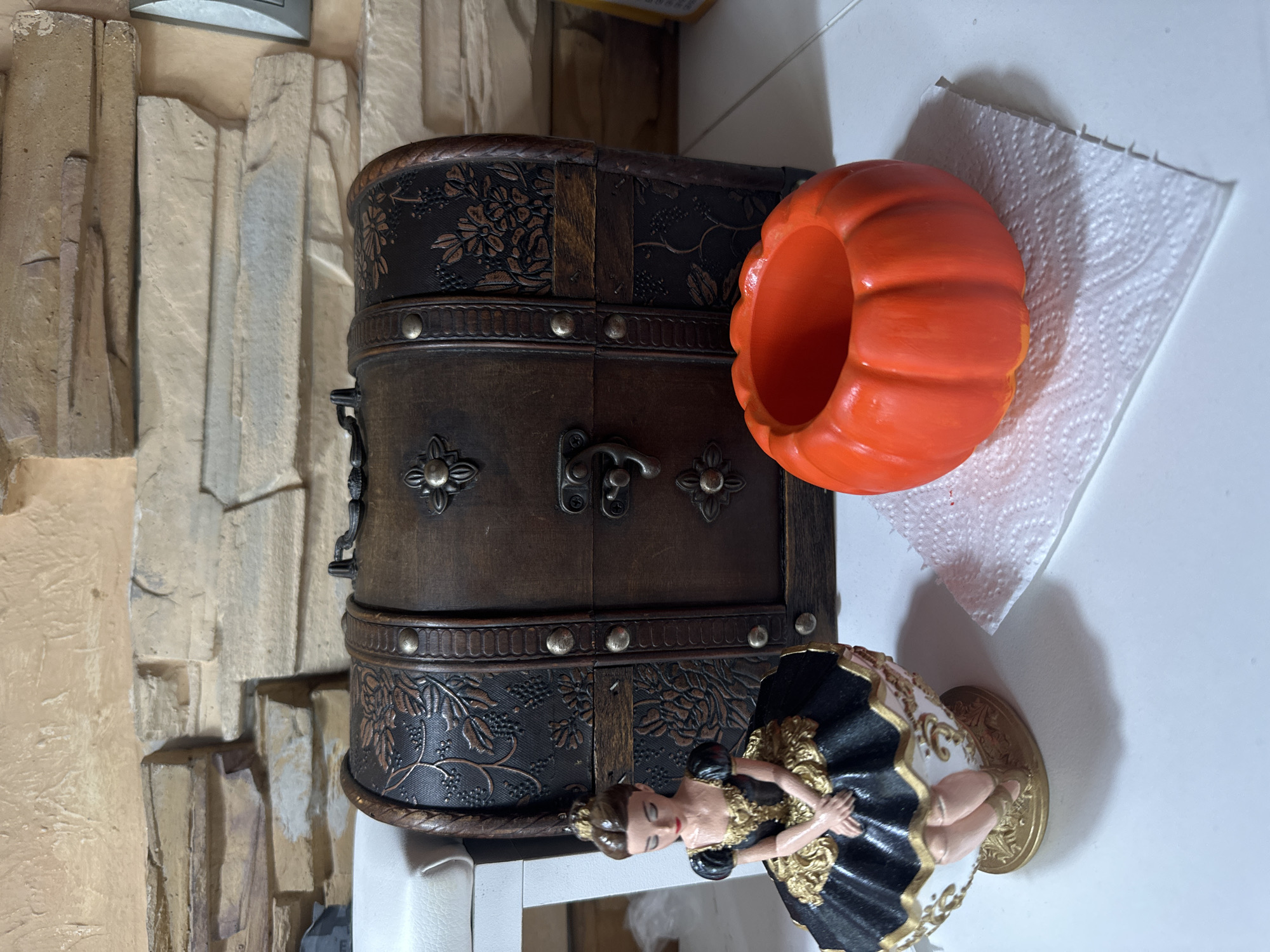 A decorative, dark brown wooden chest with embossed floral patterns and metal accents sits on a white surface. To its left is a figurine of a ballerina in a black and gold tutu, seated on an ornate base. To the right of the chest, on a white paper towel, is a small, bright orange ceramic pumpkin-shaped holder. The background consists of a textured wall with stacked rectangular stone-like elements. The image does not contain any information about the city of Vladivostok, Russia.