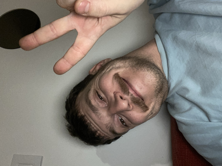 A bearded man in a light blue shirt smiles wryly, his head tilted to the right. He makes a peace sign with his fingers toward the top left. The background includes a white wall, a dark circle (likely a speaker), and a light switch. A red surface is partially visible in the bottom right. The image appears to be a selfie taken at an unusual angle, making the man look as though he is hanging upside down.