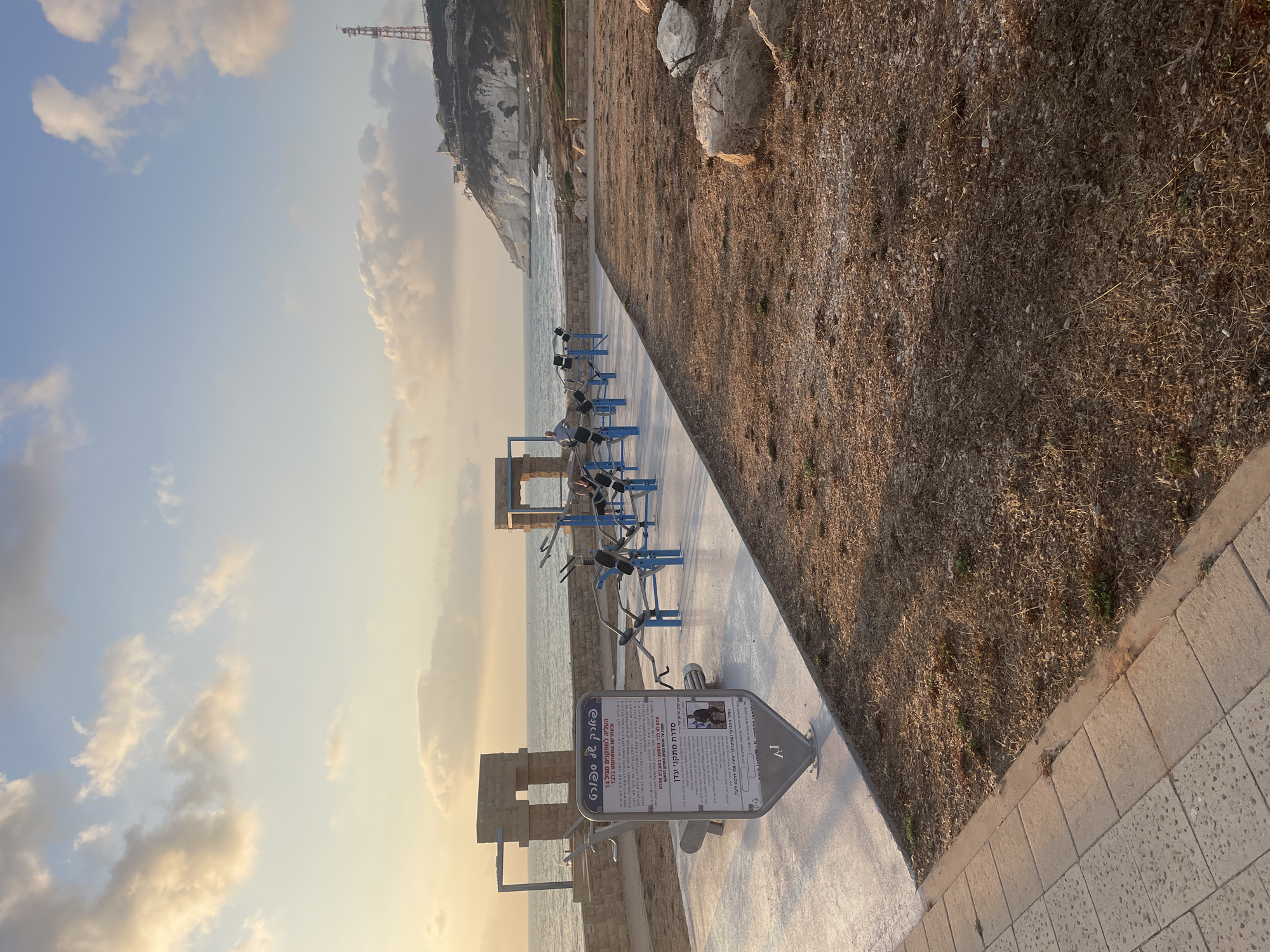 An outdoor exercise area with multiple weight machines is situated along a coastline in the Mate Asher Regional Council, Israel. The sun is low in the sky, casting a warm glow and long shadows. The sea is calm and stretches to the horizon, meeting a sky filled with soft, golden-tinged clouds. In the background, a steep, white cliff face rises above the water, topped with a telecommunications tower and structures. The exercise area features a paved walkway with several blue and silver weight machines arranged in a row. A large informational sign with text in Hebrew is prominently displayed in the foreground. Adjacent to the paved area is a patch of dry, brown grass with scattered rocks. The overall scene depicts a public space designed for recreation and relaxation by the sea.
