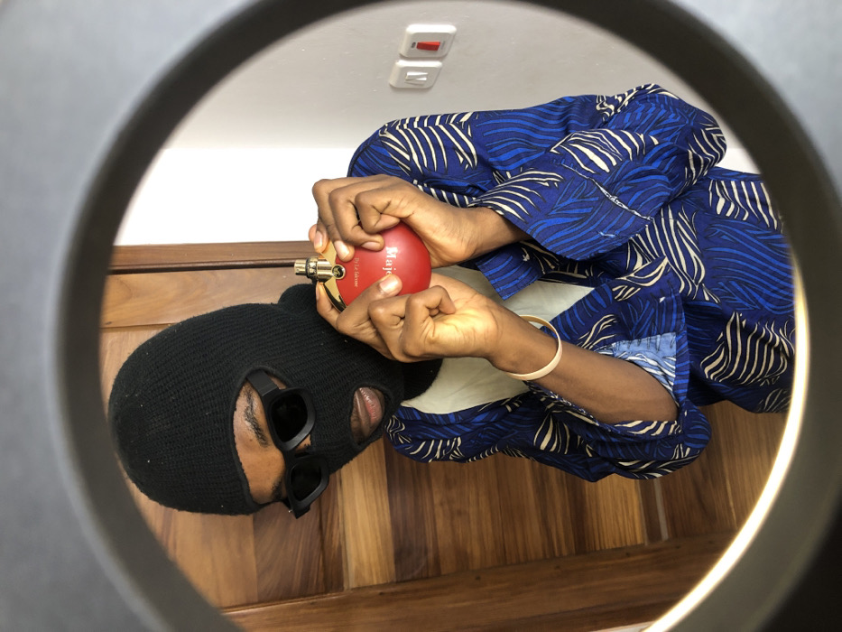 An individual is depicted within a circular photographic frame, facing forward. The person wears a black knit balaclava covering the head and most of the face, with openings for eyes and mouth. Black rectangular sunglasses obscure the eyes. The subject wears a blue and white patterned long-sleeved garment with a light-colored collar. A light-colored bangle is present on the subject's right wrist. Both hands grasp a spherical red perfume bottle, which features a gold-colored sprayer and is labeled "Majestic by Le Faisan." The background on the left consists of a light brown wooden door or panel, while the right side shows a plain white wall with two electrical switches, one of which includes a red component. The scene is located in Tema, Ghana.