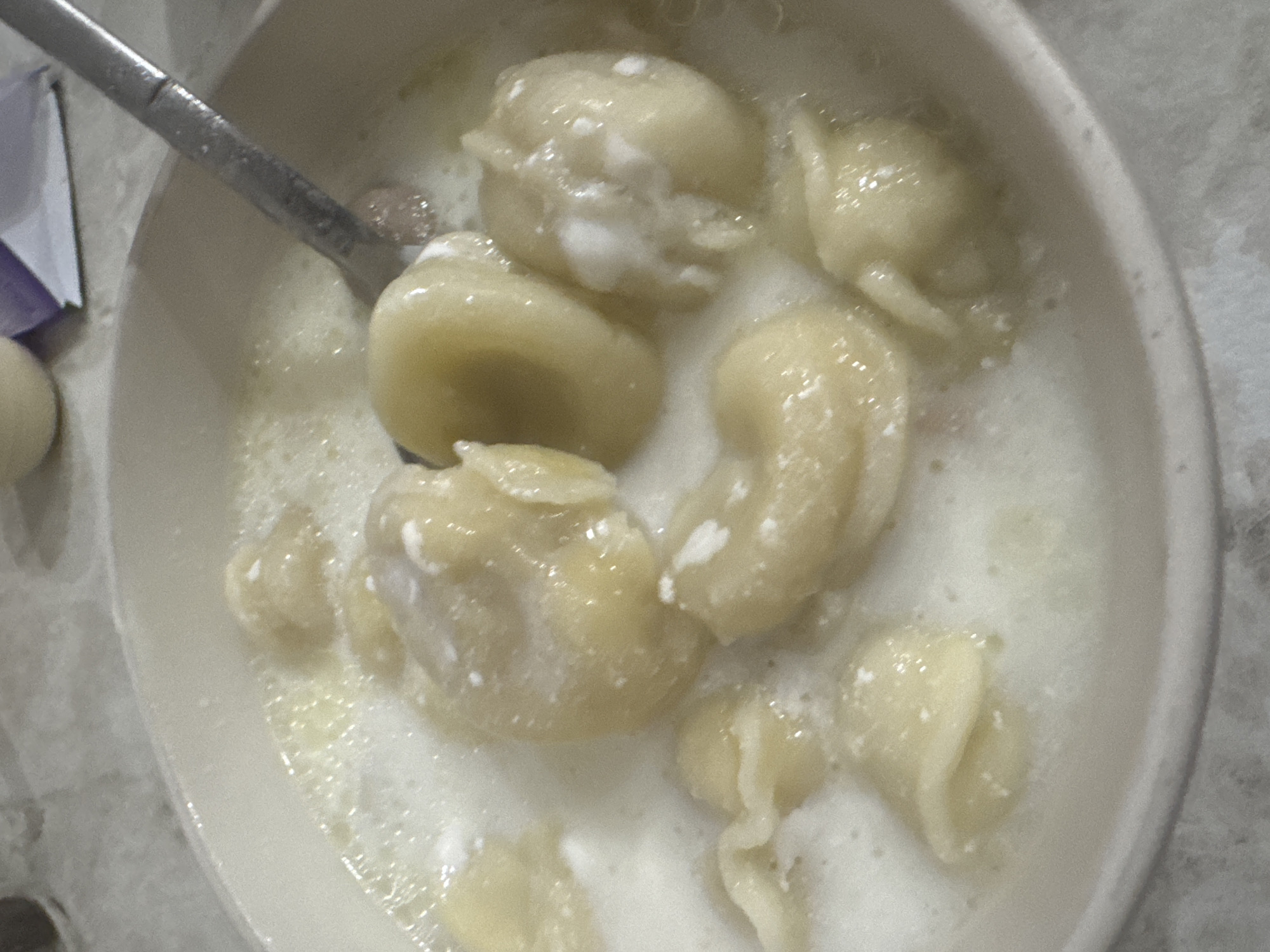 The image shows a close-up view of a bowl of food, likely pierogi or dumplings, submerged in a creamy white liquid, possibly sour cream or a milk-based sauce. A metal spoon is visible in the upper right portion of the bowl, resting partially in the food. The food items are golden yellow and appear to be somewhat soft. The bowl is a light beige color, and the surface it rests on appears to be a light-colored, speckled countertop. The background is out of focus and shows hints of a purple and white package. There are no people or animals visible in the image. The scene is indoors, likely in a kitchen or dining area. There is no information in the image to suggest a specific location in Saratov, Russia.