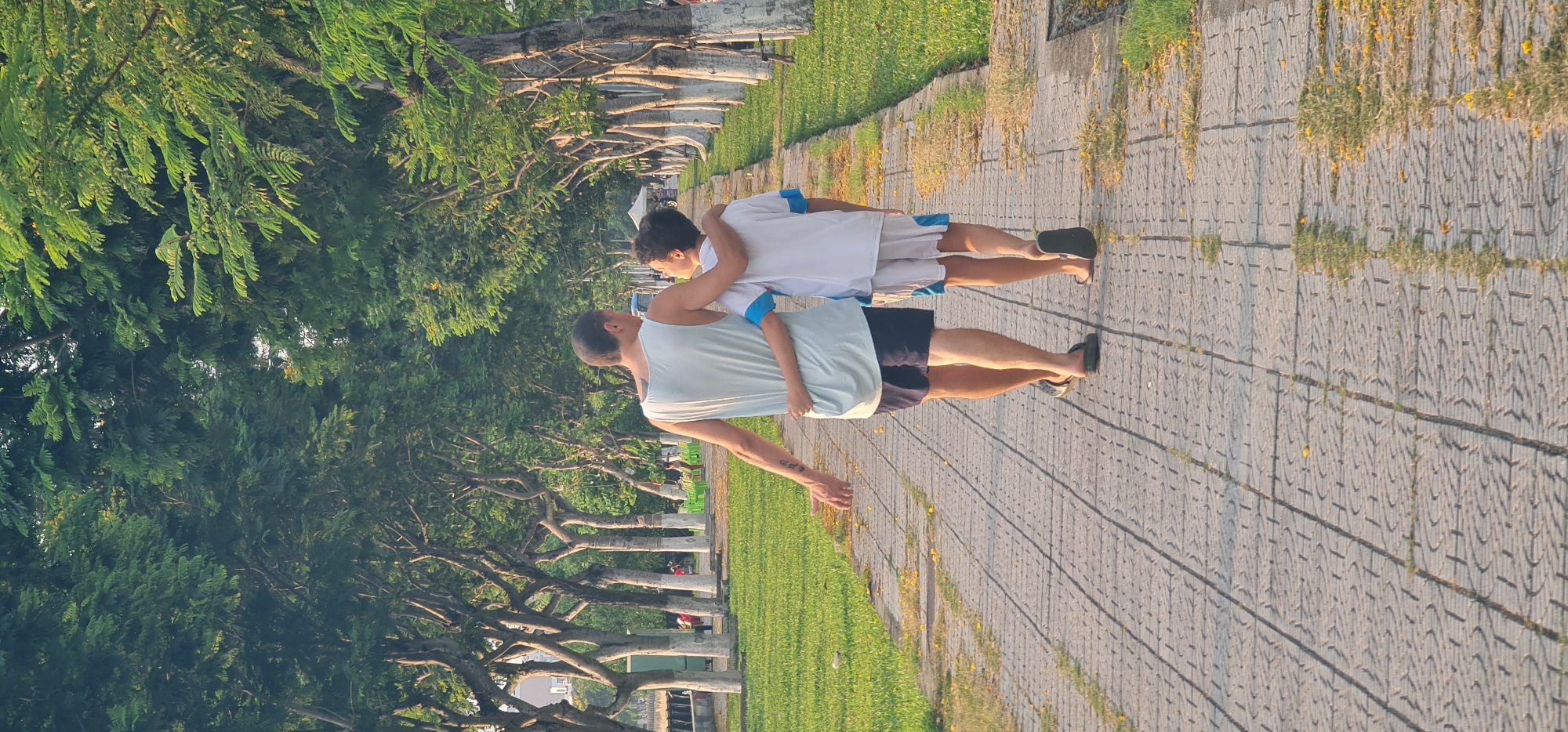 An outdoor scene captures two individuals, an adult male and a younger person, walking away from the camera along a paved park path. The adult, positioned in the foreground, wears a light blue sleeveless top and dark shorts, with a tattoo visible on his left arm. The younger person, slightly behind and to the right, is clad in a white t-shirt with blue accents on the sleeves and white shorts, with an arm gently wrapped around the adult's waist or back, suggesting a close relationship or affectionate gesture. Both are wearing dark-colored sandals or flip-flops.
The path beneath them is made of grey interlocking concrete blocks, bordered by green grass patches and scattered small yellow flowers. A distinct avenue of mature trees, featuring visible gnarled trunks and lush green foliage, lines both sides of the walkway, extending into the background.
The lighting indicates either late afternoon or early morning, characterized by a warm, golden hue that casts long shadows across the path. The clear weather suggests a pleasant day for a stroll. In the distant background, faint structures and further greenery are visible, hinting at an urban park setting, consistent with a location like Đà Nẵng, Vietnam. The overall mood is serene and peaceful, highlighting a moment of companionship. There is no visible text in the image.