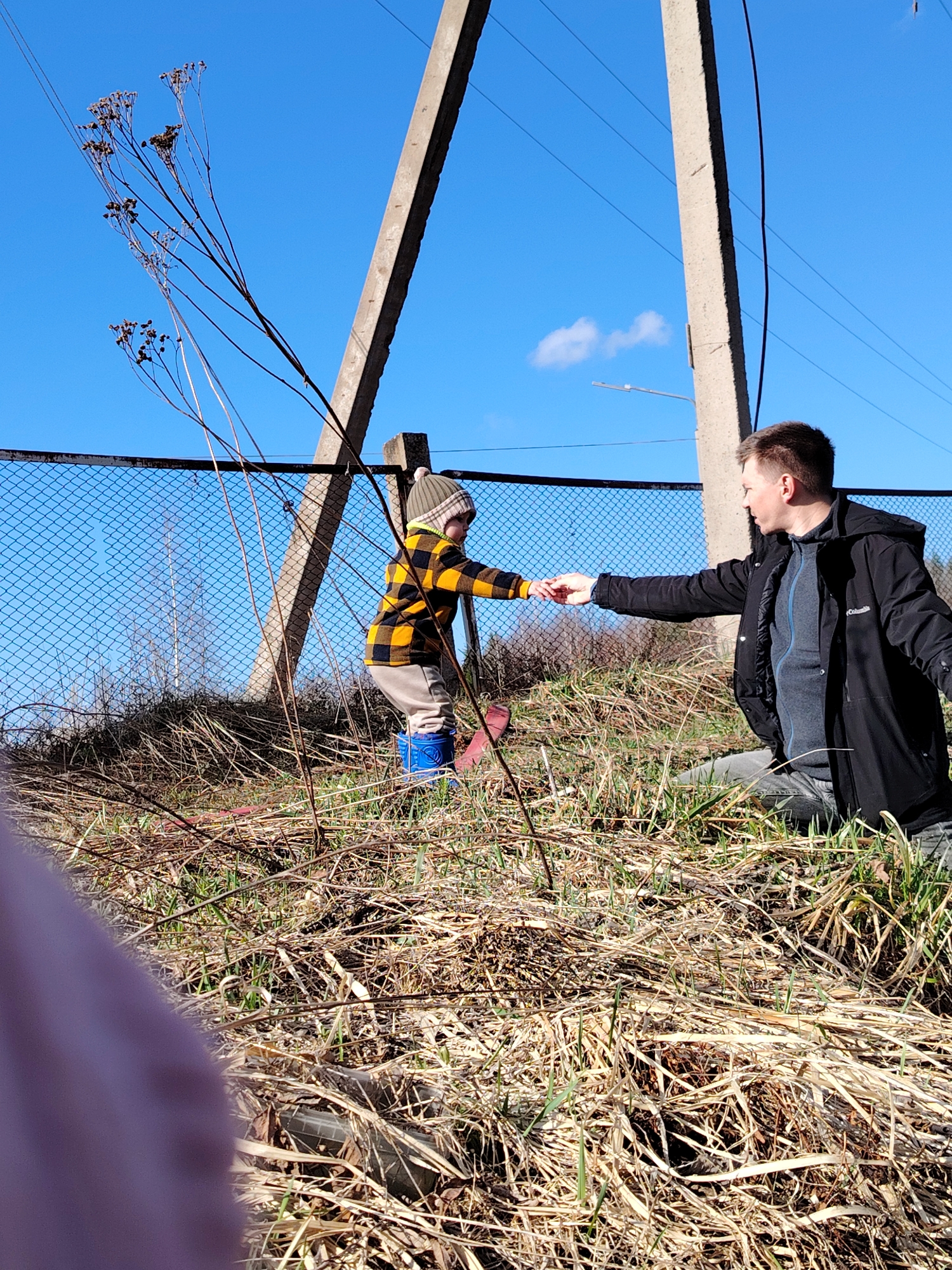 A man assists a young child while they are outside near a chain-link fence in Chaikovsky, Russia.
The man, wearing a black jacket over a gray fleece and gray pants, is kneeling on the ground, extending his right hand to the child. The child, dressed in a yellow and black plaid shirt, tan pants, a striped hat, and blue rubber boots, is reaching out to take the man's hand. The ground is covered with dry grass and some green patches. Behind the fence, there are trees and utility poles against a clear blue sky with a single small white cloud. Tall, dry weeds are in the foreground. A portion of a purple object is present in the lower-left corner of the frame.