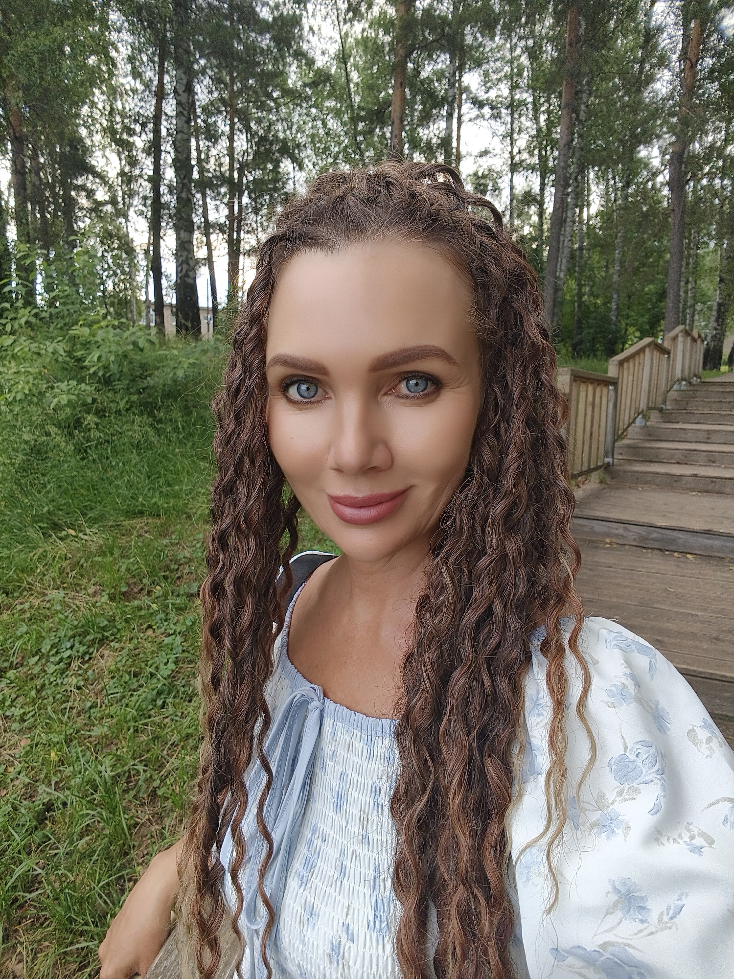 A woman with curly brown hair and blue eyes is taking a selfie outdoors. She is wearing a white dress with a blue floral pattern. Behind her, a wooden staircase leads up a grassy slope with trees. The location is the Chaikovsky Nature Reserve in Chaikovsky, Russia.