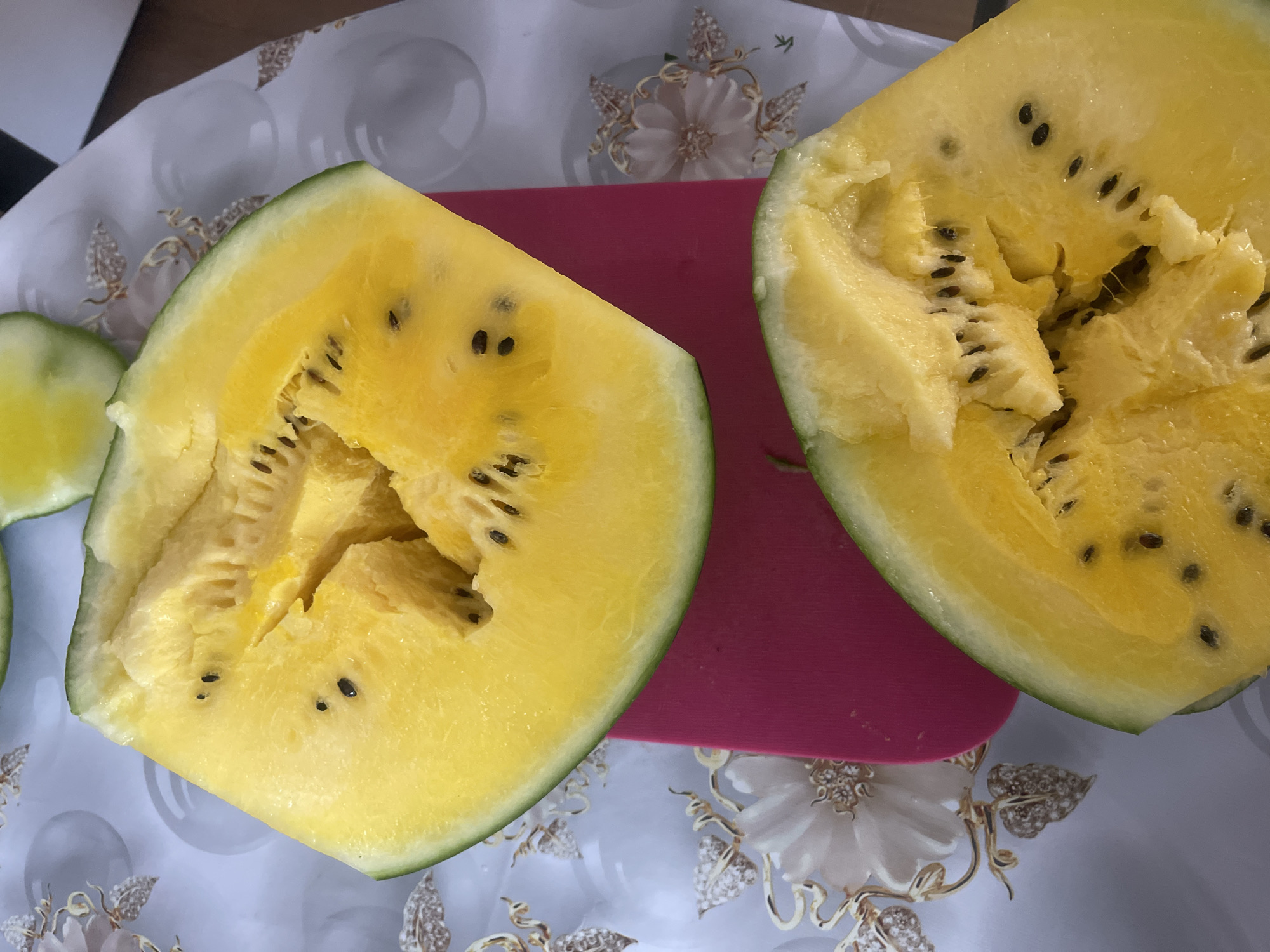 Two halves of a yellow watermelon are displayed on a patterned tablecloth. One half is positioned at the top of the image and the other at the bottom. A bright pink cutting board is partially visible beneath the top watermelon half. The watermelon flesh is a vibrant yellow with numerous small, black seeds scattered throughout. The rind is a pale green. The background shows a white tablecloth with a decorative floral pattern in gold and light pink. There are no people or animals visible in the image. The scene appears to be indoors, likely in a kitchen or dining area.