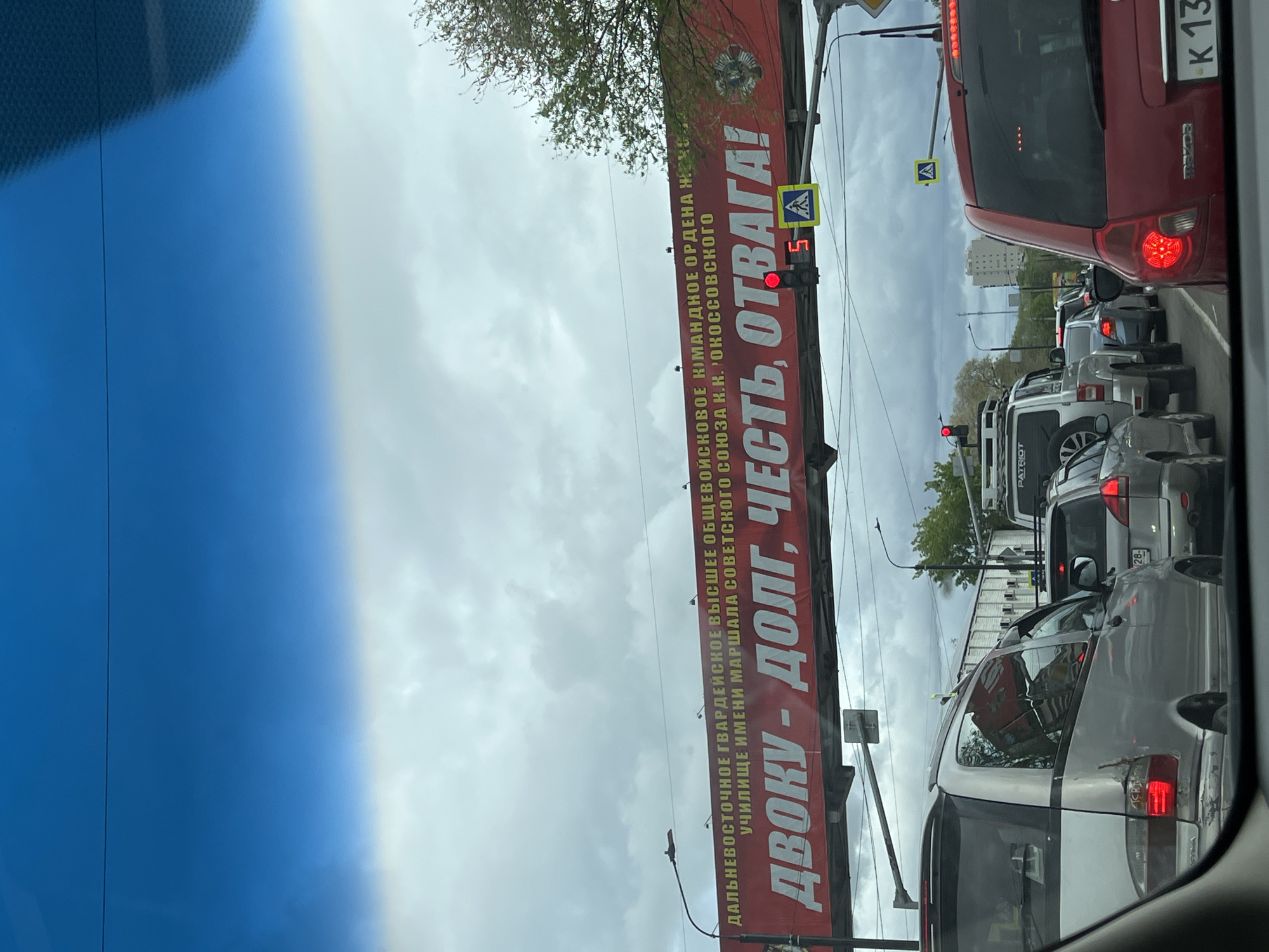 The image shows a street scene in an unknown city, viewed from inside a car. The sky is cloudy. Traffic is stopped, with several cars visible in front of the viewer's vehicle. A large, red banner hangs across the street. The banner reads, "ДАЛЬНЕВОСТОЧНОЕ ГВАРДЕЙСКОЕ ВЫСШЕЕ ОБЩЕВОЙСКОВОЕ КОМАНДНОЕ ОРДЕНА Ж УЧИЛИЩЕ ИМЕНИ МАРШАЛА СОВЕТСКОГО СОЮЗА К.К. РОКОССОВСКОГО ДВОКУ - ДОЛГ, ЧЕСТЬ, ОТВАГА!" (Far Eastern Guards Higher Military Command Order of J School named after Marshal of the Soviet Union K.K. Rokossovsky DVOKU - Duty, Honor, Courage!). A traffic light shows a red light with a countdown of 5. A pedestrian crossing sign is visible near the traffic light. A license plate, "K 13", can be seen on the back of the car in front.