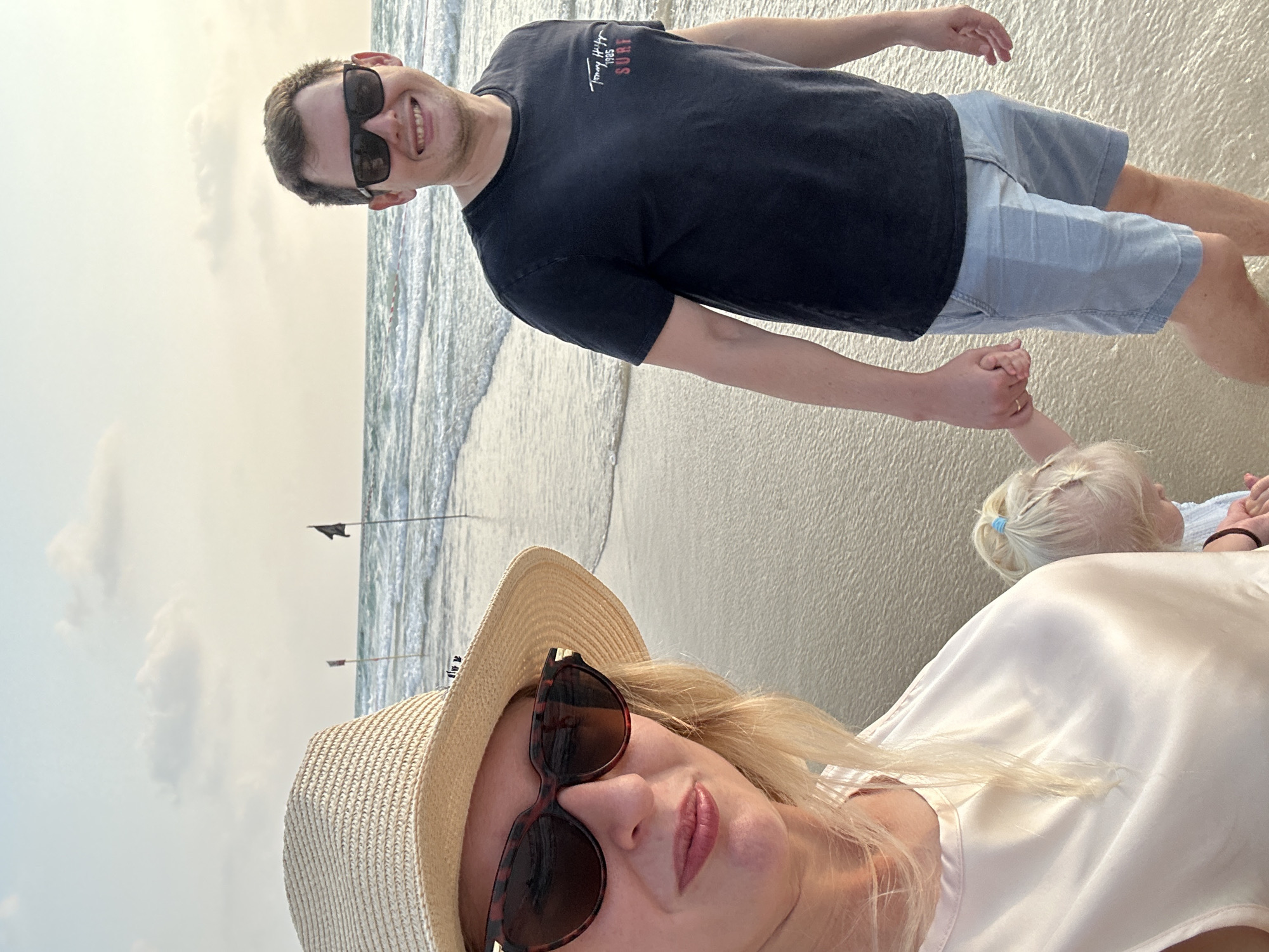 The image depicts a family on the beach in Bat Yam, Israel. A woman wearing a straw hat and sunglasses is in the foreground, on the left, facing the camera. To her right, a man wearing sunglasses and a dark t-shirt and light blue shorts is also facing the camera, smiling. The man is holding the hand of a young child whose back is to the camera. The child has blonde hair tied with a blue ribbon. The family is standing on the wet sand near the waterline, with gentle waves in the background. Two black flags on poles are visible in the distance on the sea. The sky is overcast with some clouds. The scene appears to be a casual outing at sunset.