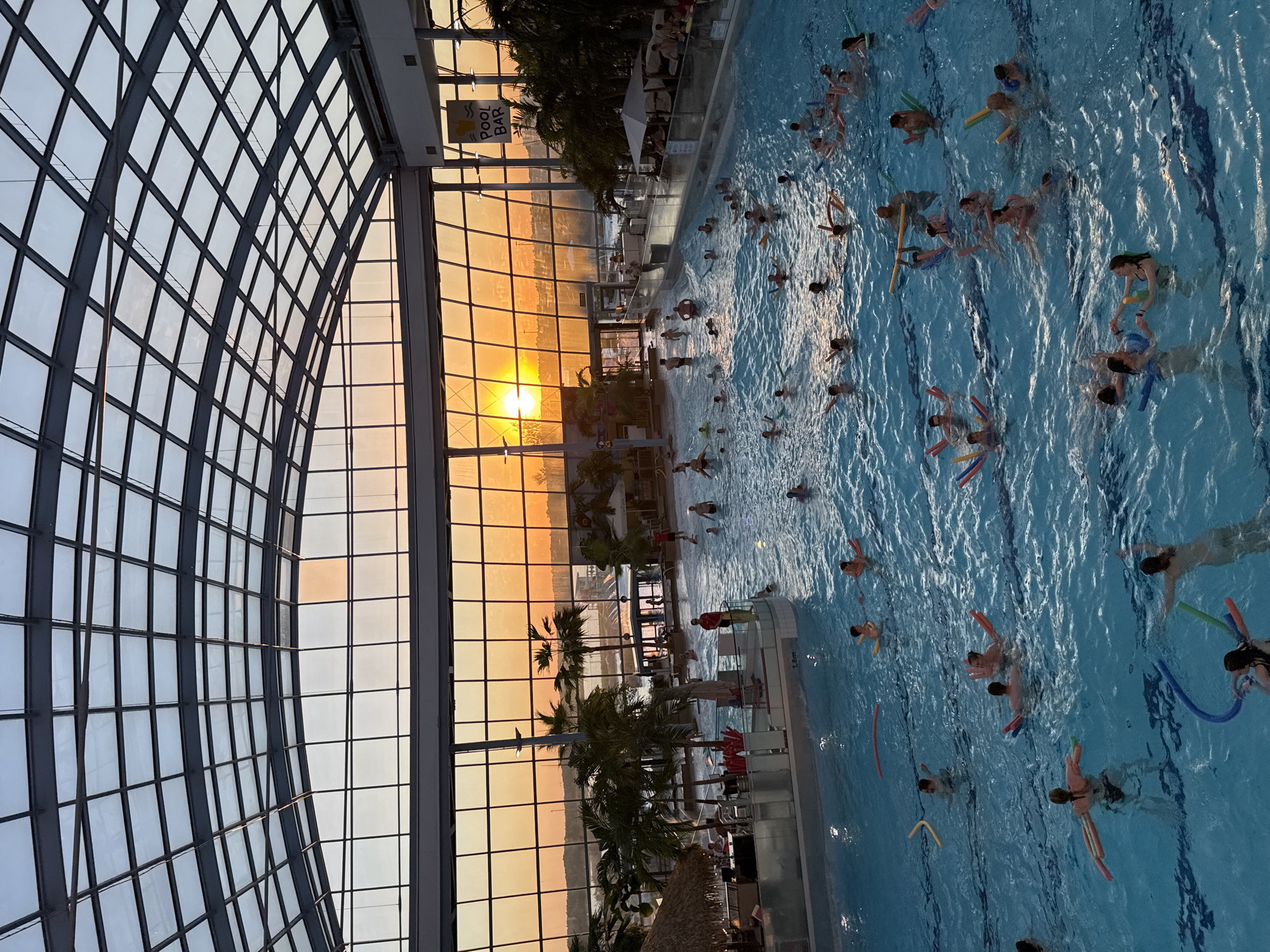 The image shows a large indoor swimming pool filled with people swimming and playing. Many of them use floating devices. The pool is situated within a building with a large glass wall, and a sunset is visible through the glass, suggesting an evening time. Palm trees are placed near the edge of the pool, giving it a tropical feel. A sign is visible, seemingly indicating a pool bar. The image presents a leisure scene, with people enjoying swimming in a controlled environment while observing the outdoor sunset. The precise location is not apparent, but it appears to be a recreational facility.