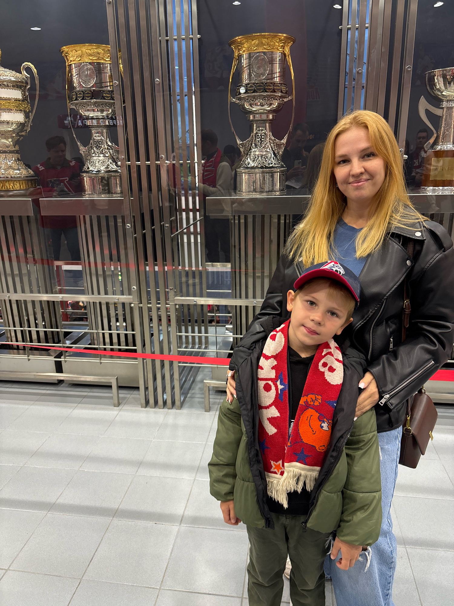 A woman with blonde hair and a black leather jacket stands next to a young boy wearing a green puffer jacket, a red and white scarf, and a baseball cap. The woman's left arm is around the boy's shoulder. They are posed in front of display cases containing large, ornate trophies. The background shows the trophies are housed in what appears to be a museum or sports hall, with metallic vertical bars and glass enclosures. The floor is tiled with light gray squares. The overall environment suggests a public space dedicated to sporting achievements.