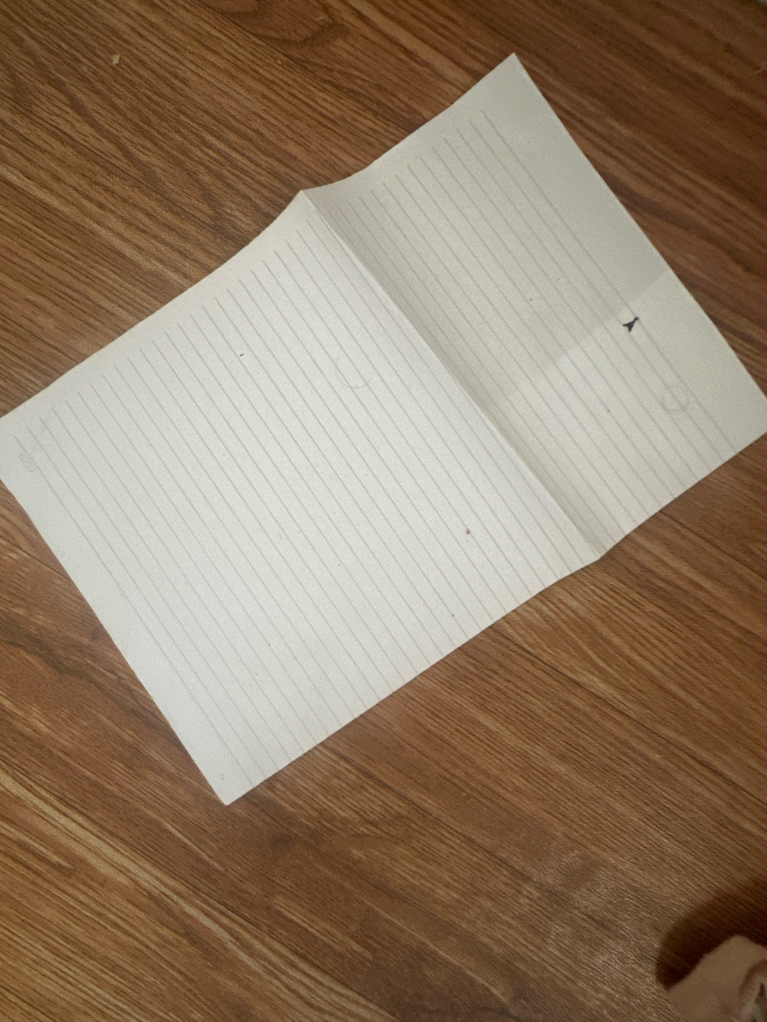 An overhead shot shows a folded piece of lined white paper resting on a brown wooden surface. The paper is positioned diagonally from the lower left to the upper right of the frame, with its left side closer to the viewer. The paper appears to be a single sheet that has been folded in half, creating a visible crease down the middle. The surface it rests on is made of wooden planks with a prominent grain pattern. The lighting casts subtle shadows and highlights across the scene. There are no people or other objects visible in the frame. The scene is devoid of any context suggesting a specific location in Margamulya, Indonesia.