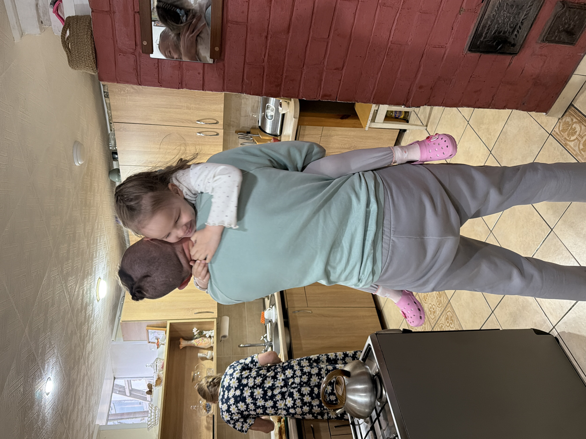 The image depicts a man carrying a young girl on his back in what appears to be a kitchen. The man is wearing a light green shirt and grey sweatpants. The girl is wearing a white top and pink shoes. An older woman in a blue and white daisy-patterned dress is standing near a stove with a kettle on it. The kitchen has light-colored cabinets and tiled floors. Based on the provided information, the location is Chaikovsky, Russia.