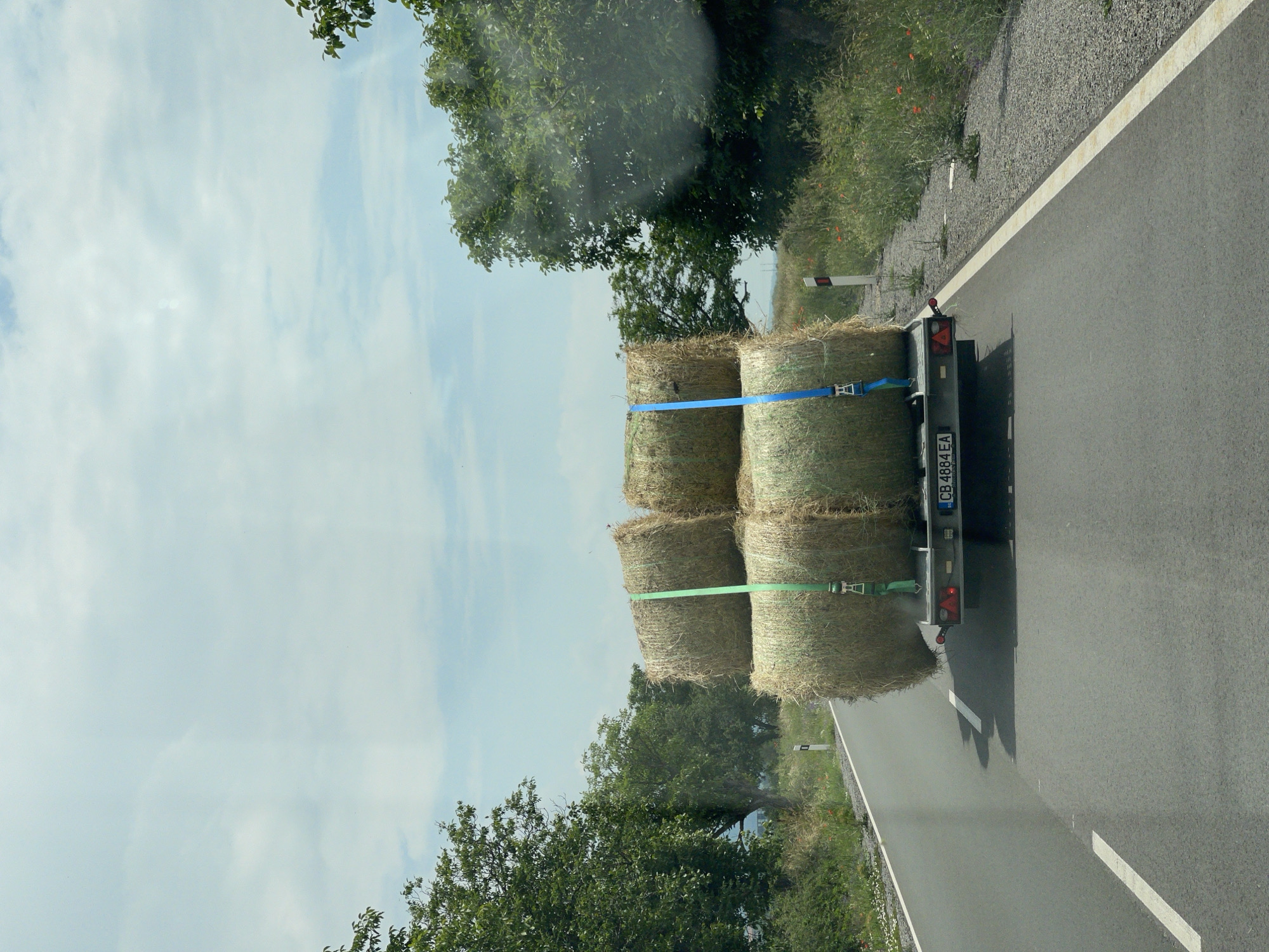 The image displays a rural road scene in Karnare, Plovdiv Province, Bulgaria, viewed from inside a vehicle looking forward. A silver-grey utility trailer is observed from the rear, traveling away on a paved asphalt road. The trailer is loaded with four large, cylindrical bales of hay, stacked two horizontally on the trailer bed and two directly on top. These bales are secured by one green and one blue ratchet strap. The trailer's rear features a Bulgarian license plate, "CB 4884 EA", and unlit red triangular reflectors and tail lights. The asphalt road has a solid white line on the right shoulder and a dashed white line on the left. Both sides of the road are flanked by gravel verges, transitioning into dense green deciduous trees and wild vegetation, including scattered red poppies. White and red reflective road marker posts are visible on both verges. The sky above is light blue with scattered white, diffuse clouds. No individuals are present in the scene.
