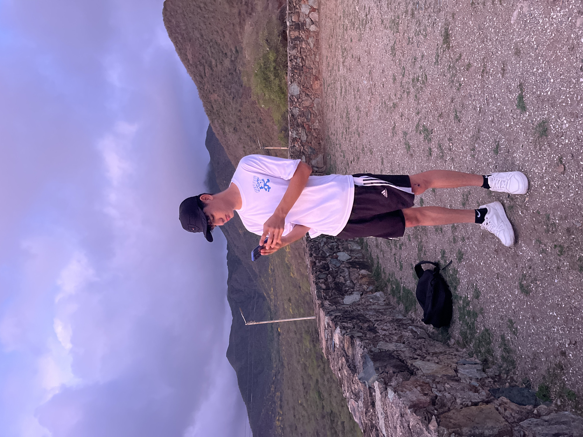 A young person wearing a black baseball cap, white t-shirt, and black athletic shorts stands outdoors, looking at their smartphone. They are wearing white sneakers and black socks. A black backpack is on the ground next to a stone wall. The background features mountainous terrain with sparse vegetation and a cloudy sky. This location is believed to be in the city of Santa Ana, Venezuela, known for its natural landscapes.