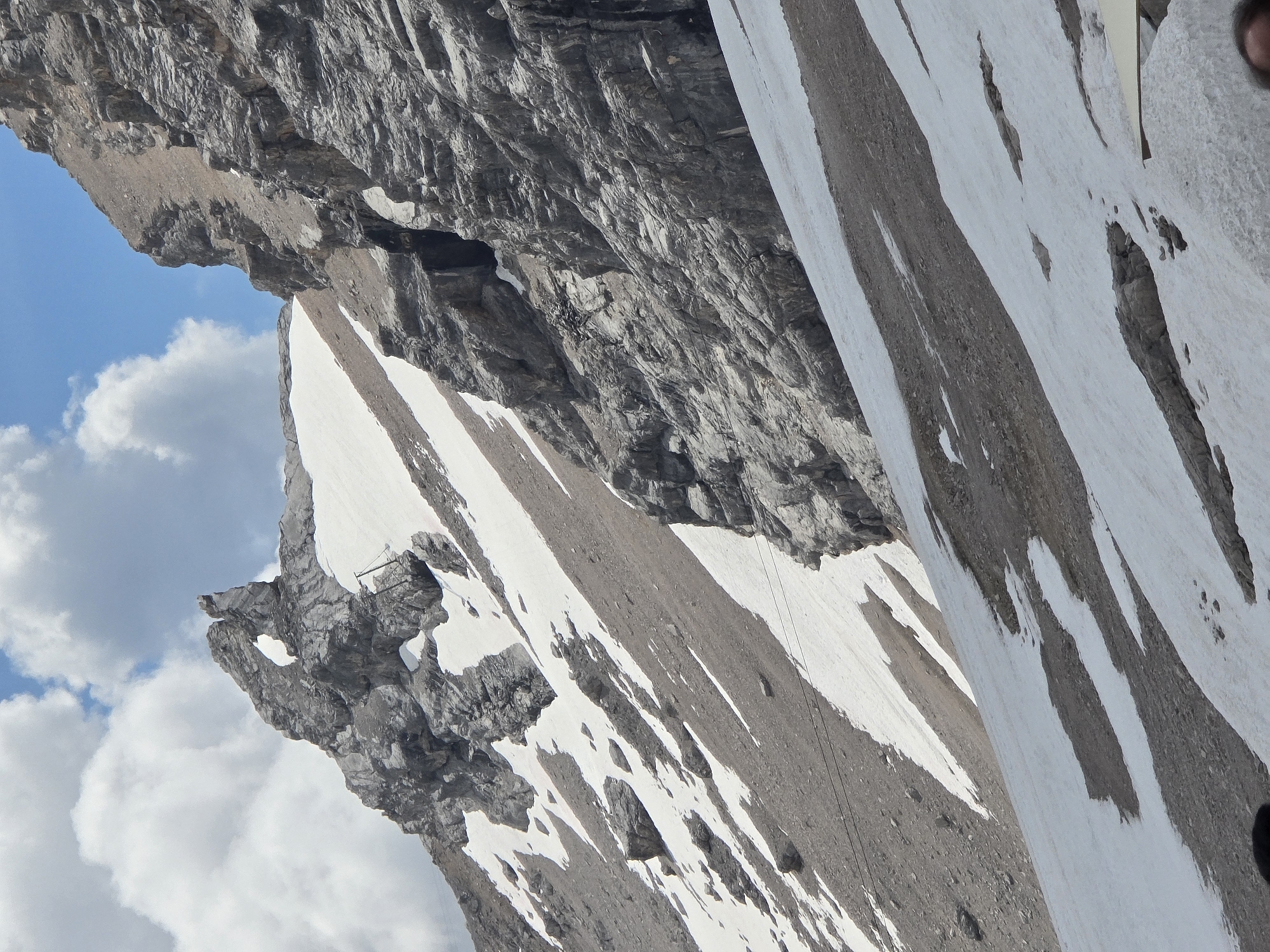 The image depicts a mountainous landscape with snow patches and rocky cliffs. A prominent rock formation with a large hole in it is visible on the left side of the image, against a blue sky with white clouds. The terrain is steep and appears to be a hiking or climbing area. There are also some cables or wires running across the slope, possibly related to a cable car or ski lift. The location is identified as the Zugspitze, the highest peak in Germany, near Garmisch-Partenkirchen. The image captures the natural beauty and ruggedness of the alpine environment.