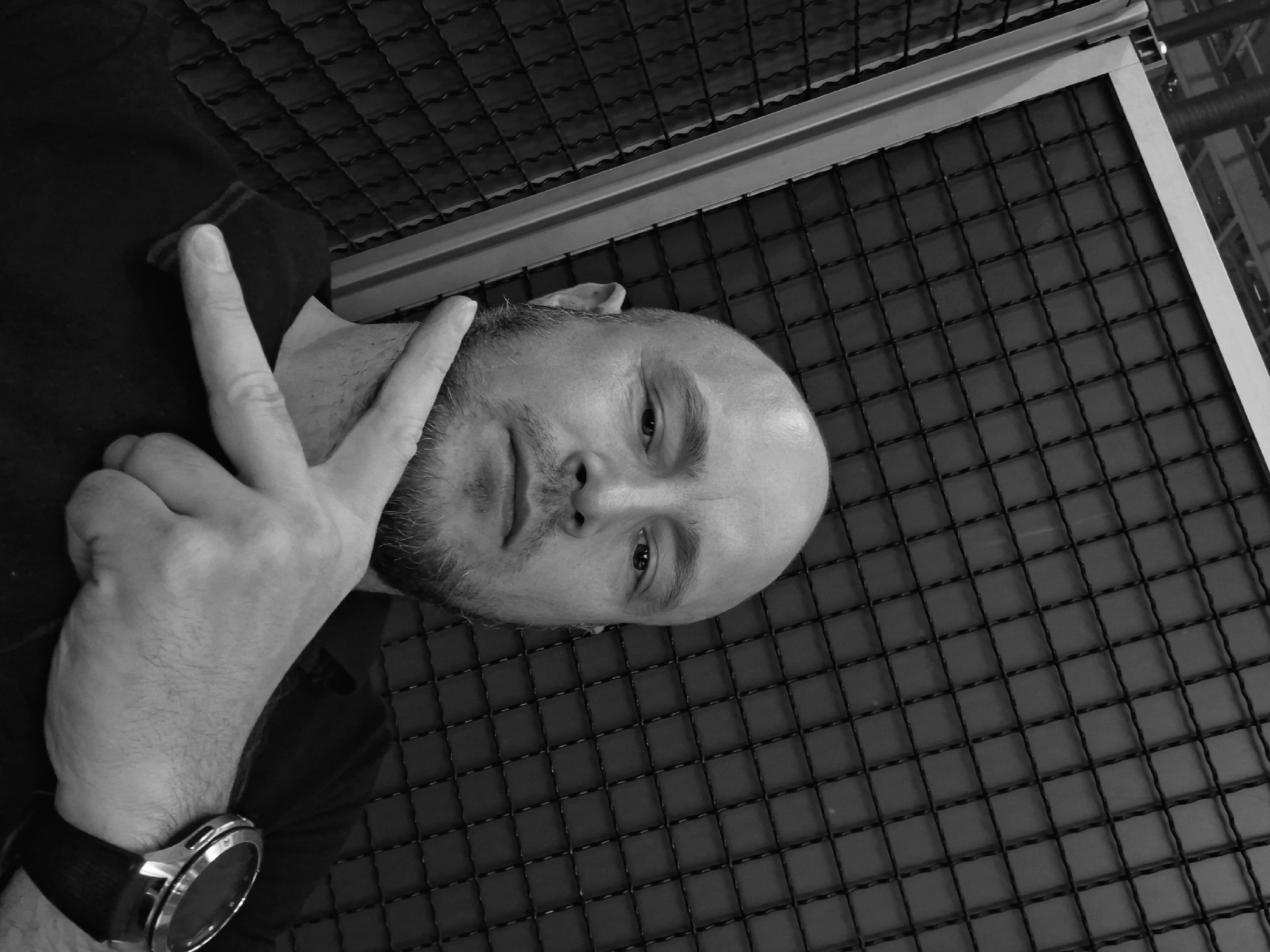 A black and white, medium shot, waist-up photograph shows a bald man with a goatee. He is looking directly at the camera and has his right index and middle fingers extended, making a peace sign. The man is wearing a dark shirt and a watch on his left wrist. He is positioned in front of a dark, grid-like textured background. The lighting is even, and the overall image has a serious tone. The location is Unknown, Unknown.
