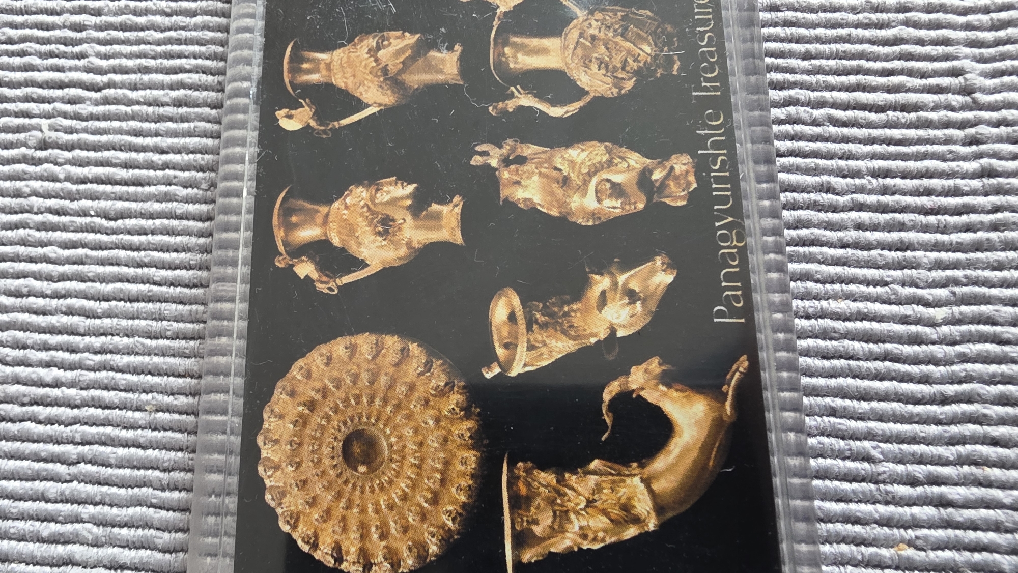 The image shows a photograph of a postcard depicting the Panagyurishte Treasure. The postcard showcases several golden artifacts, including ornate vessels and rhytons, arranged on a black background. Text on the card reads "Panagyurishte Treasure". The postcard is encased in a clear plastic sleeve and is placed on a gray textured surface. The Panagyurishte Treasure was found in Panagyurishte, Bulgaria, not in Blagoevgrad.