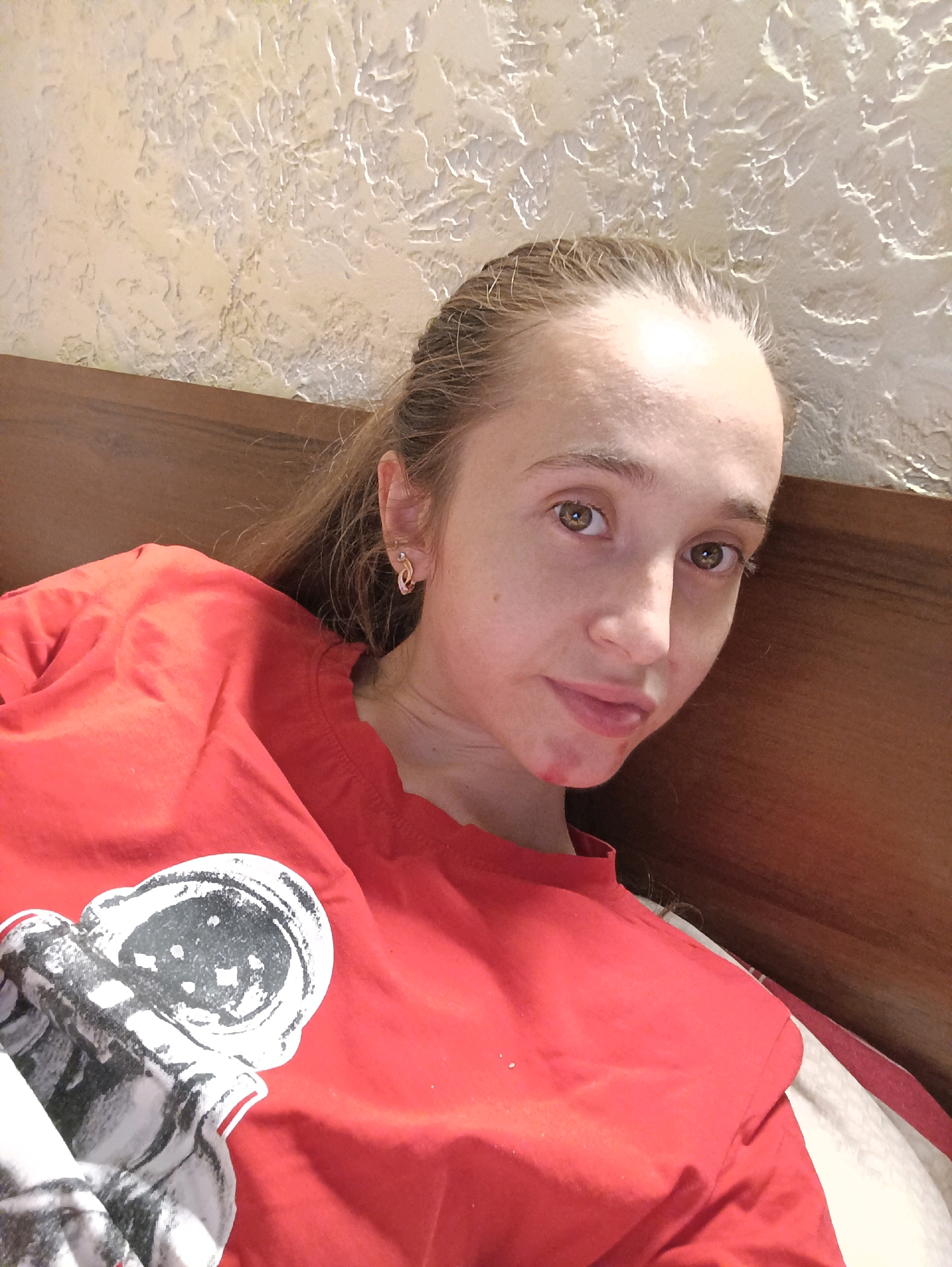 A young woman with long, light brown hair pulled back is lying down and looking at the camera. She is wearing a bright red t-shirt with a black and white graphic of an astronaut's helmet on the left side. The background appears to be a textured wall and a wooden headboard, suggesting she is in a bedroom. The woman's skin is pale, and she has brown eyes. There is a red mark around her mouth. The scene is lit by an overhead light source, creating soft shadows. There is no indication of a specific location within the city of Vypasne, Ukraine.
