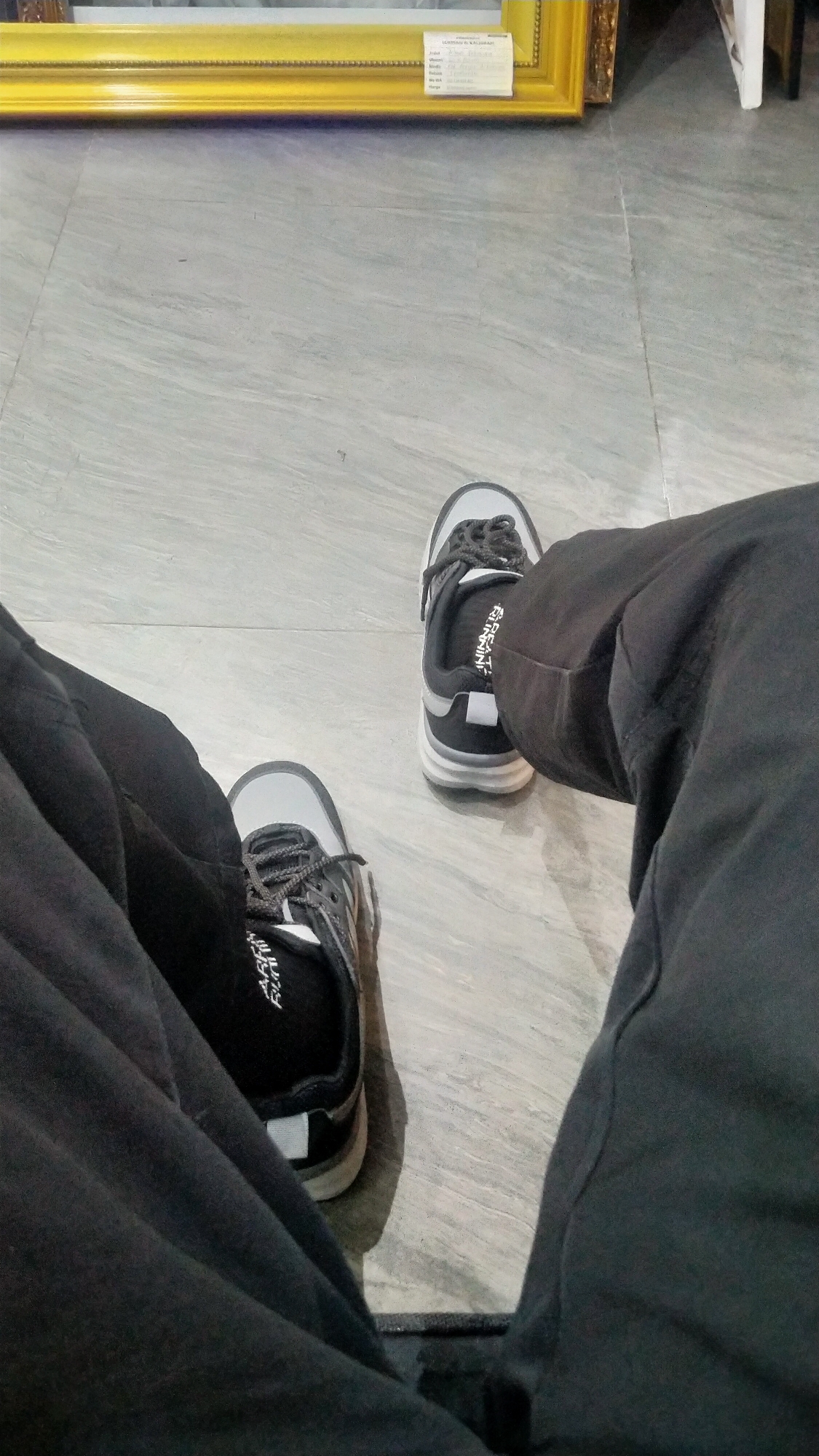 The image shows a low-angle shot of a person's legs and feet, clad in dark pants and black and gray athletic shoes, resting on a light gray tiled floor. A framed artwork with a gold frame is visible in the background, partially blurred. The artwork appears to be a print or painting with text on a white label attached to its lower right corner. The floor tiles have a marbled pattern. The scene is indoors, with a neutral, somewhat sterile ambiance. The location is specified as Nyamat, Indonesia.