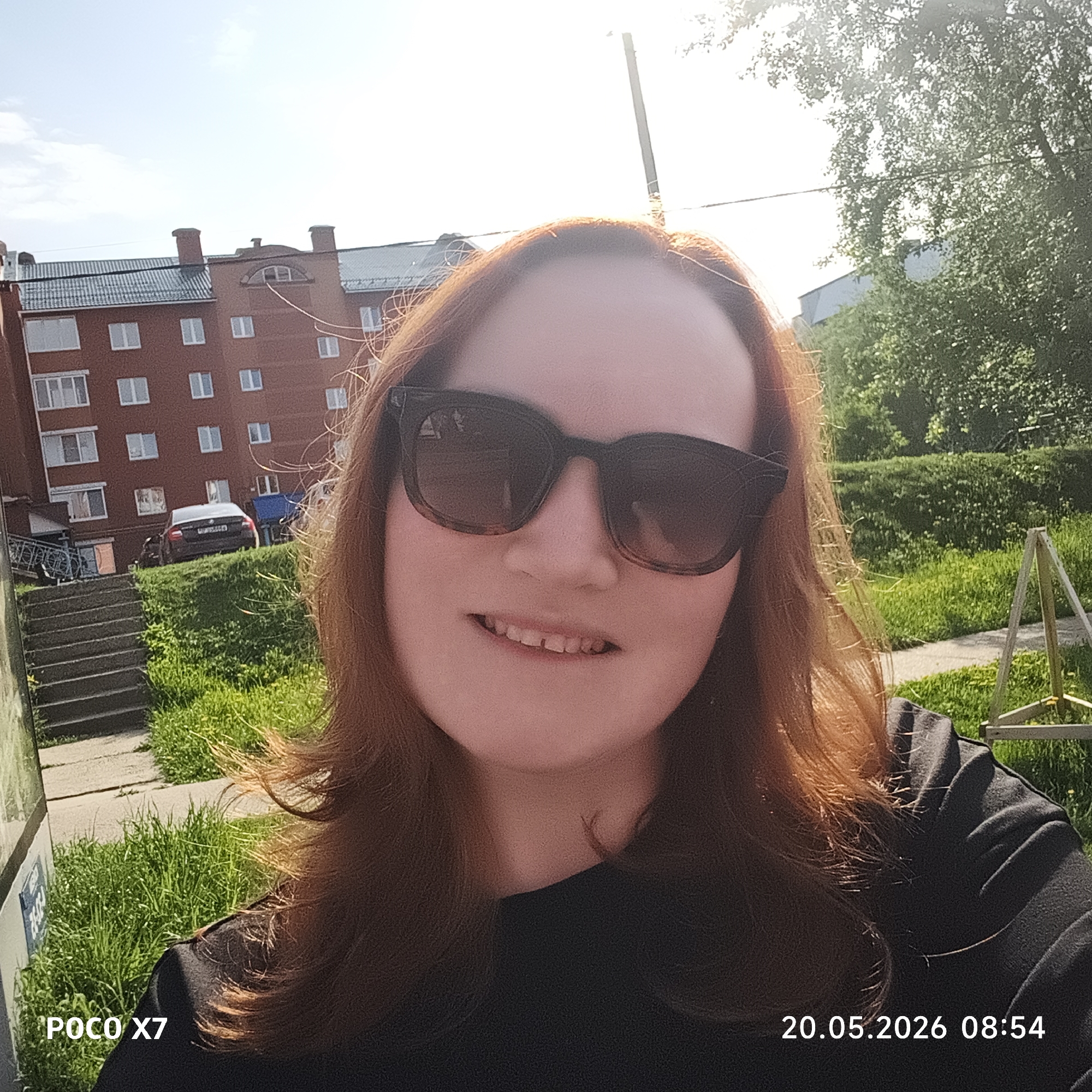 The image is a selfie of a woman wearing sunglasses, taken outdoors on a sunny day. In the background, there is a red brick building with white-framed windows. To the left of the building are concrete steps, and a dark-colored car is parked on a grassy area behind the steps. Lush green grass and some type of play equipment are visible in the mid-ground. A tree and additional foliage are on the right. The date "20.05.2026 08:54" is printed in the lower right corner. The camera logo "POCO X7" is in the lower left corner. The location is likely Chaikovsky, Russia based on the building style.