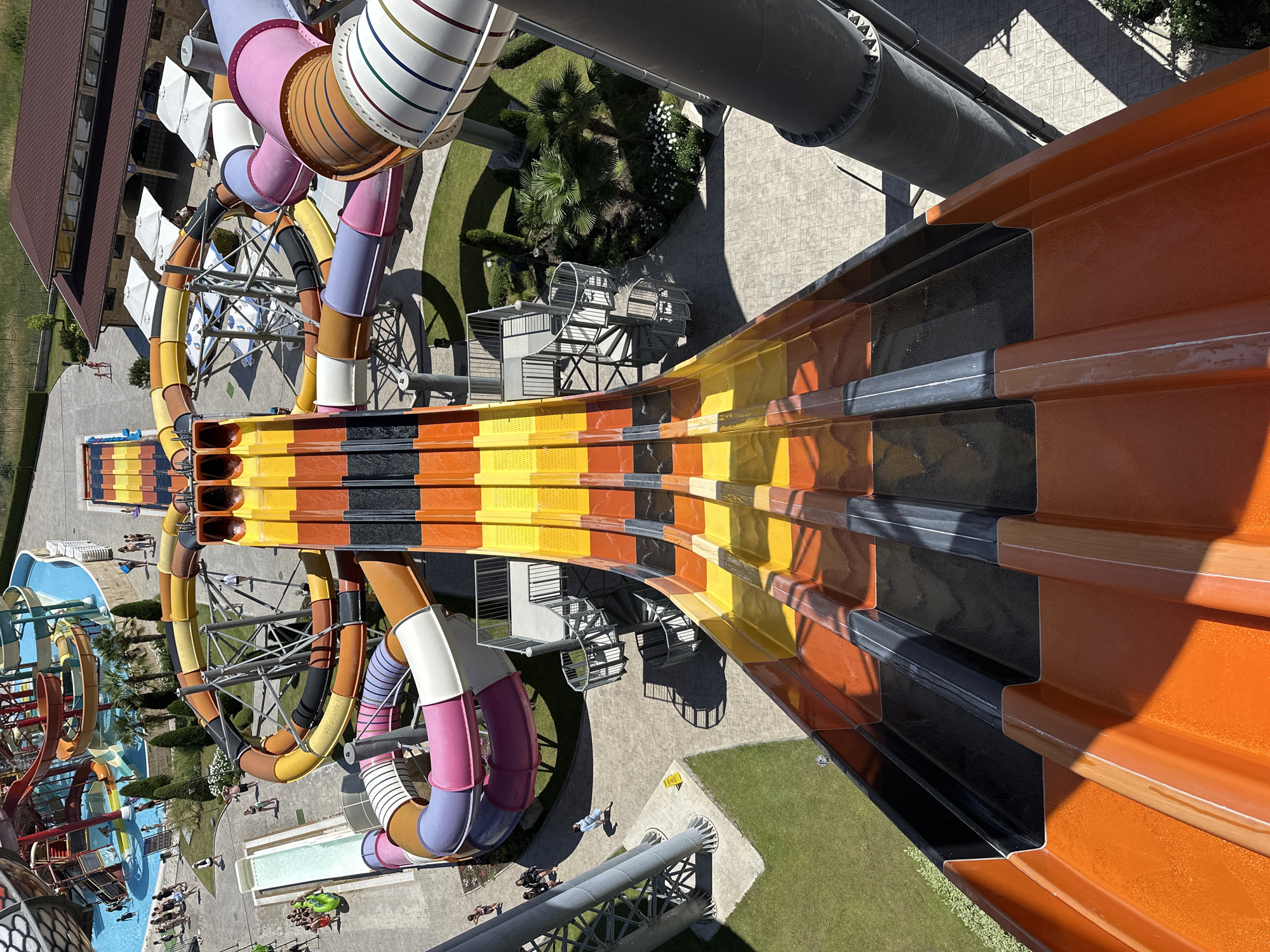 This aerial shot captures a vibrant water park on a sunny day. The main subjects are the colorful water slides, with prominent orange, yellow, black, and brown sections dominating the foreground and midground. These slides twist and turn, forming an intricate network above a green lawn and paved pathways.
In the background, a large building with a reddish-brown roof is visible. There are several people scattered throughout the park, some appearing to be waiting for the slides or relaxing by the pools. Children are visible in the pools, indicating a family-friendly atmosphere. The scene is set in an outdoor amusement park.
The activity taking place is the operation of a water park, with the slides clearly designed for amusement and recreation. The time of day appears to be daytime, likely late morning or early afternoon, given the bright sunlight and distinct shadows cast by the structures and trees. The weather seems to be clear and warm, suitable for a day at the water park.
Notable details include the metallic supports for the slides, the clean blue water in the pools, and the lush green grass. The slides themselves are a key feature, showcasing a modern design with distinct color blocks. The overall impression is one of fun and excitement. No visible text is apparent in the image. The location context of Ravda, Bulgaria, suggests a popular tourist destination.