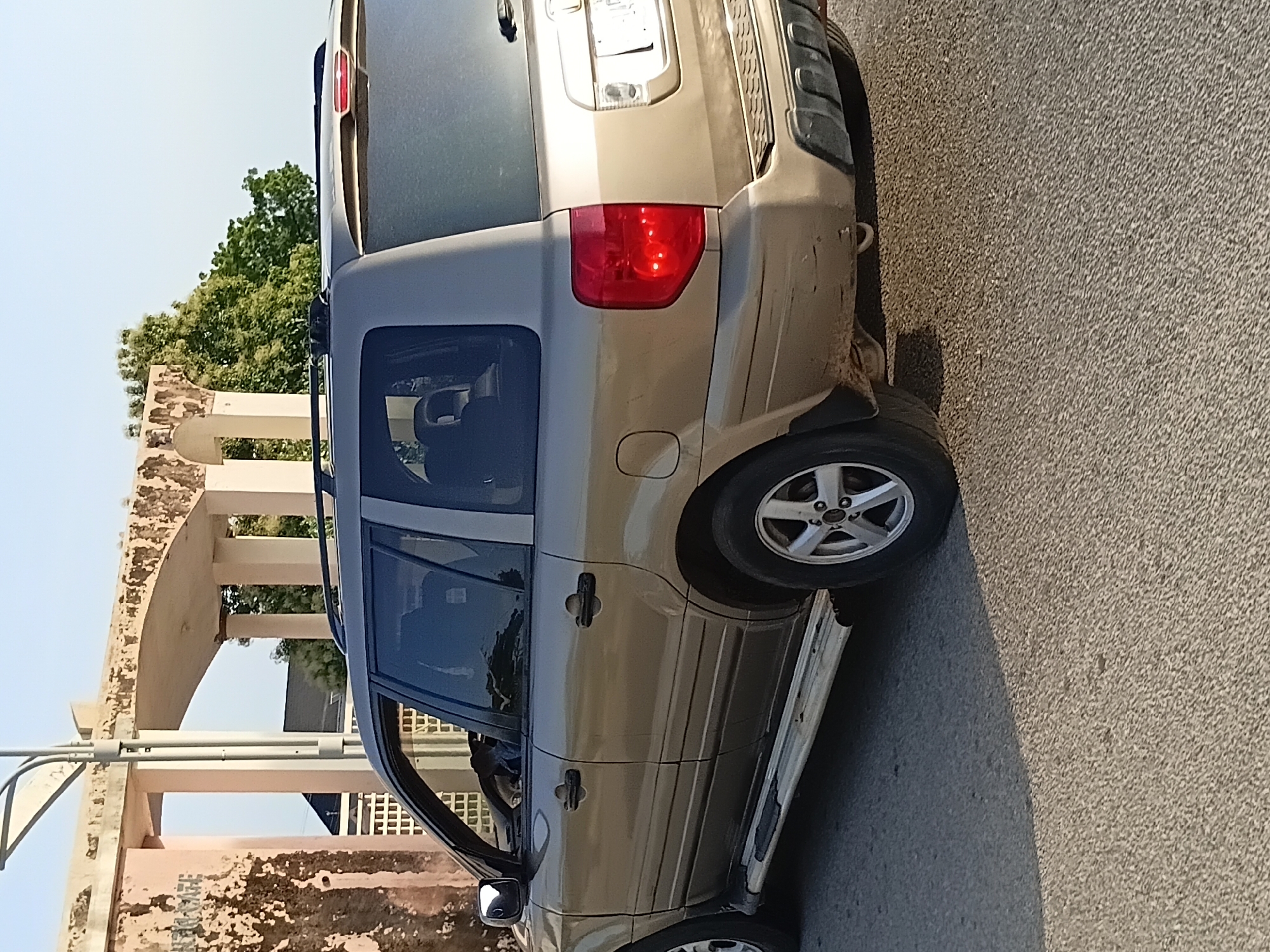 The image shows a tan-colored Honda Pilot SUV parked on a paved road in Jalingo, Nigeria. The SUV is viewed from the rear-right side. The vehicle has a roof rack and a red taillight on the right side. Behind the vehicle, there is an arched structure with weathered pillars and a textured wall. Trees with green foliage are visible beyond the structure. The sky is clear and light blue. The road surface is asphalt.