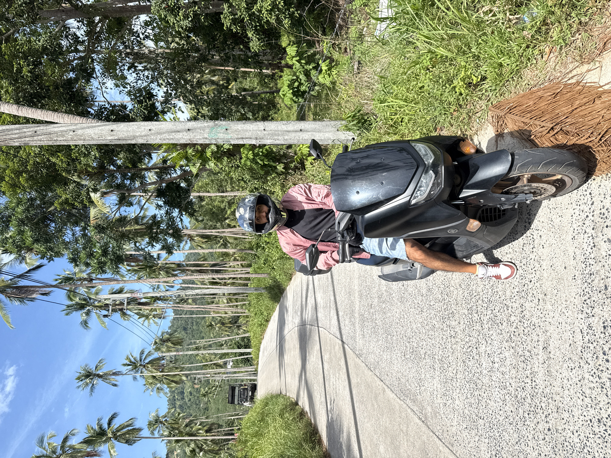 A person wearing a pink jacket, black t-shirt, denim shorts, sneakers, and a helmet is seated on a black scooter on a paved road. The road curves to the left and is flanked by lush green grass and numerous palm trees. In the background, a dark truck is visible on the road. Power lines are strung between utility poles that run along the side of the road, with a backdrop of a hilly, tree-covered landscape under a blue sky with scattered clouds. The scene appears to be located on the island of Ko Samui in Thailand, a tropical destination known for its beaches, coconut plantations, and scenic roads.
