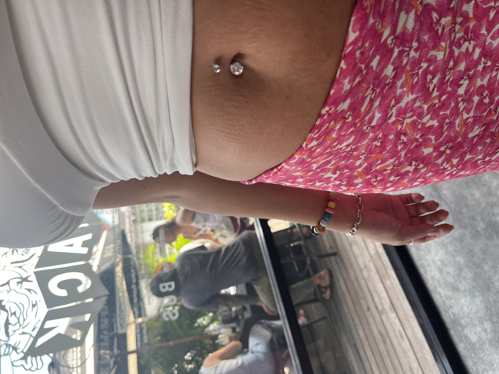 A close-up shot focuses on a person's midriff, revealing a belly button piercing with two silver studs. The individual is wearing a white crop top and a pink floral skirt. Their arm, adorned with a colorful beaded bracelet and a silver chain bracelet, is visible on the right side of the frame. In the blurred background, through a window, patrons of an establishment can be seen seated, with a sign reading "BACK" partially visible on the left. The setting appears to be a cafe or restaurant in North Kuta, Indonesia, with wooden flooring and greenery visible outside.