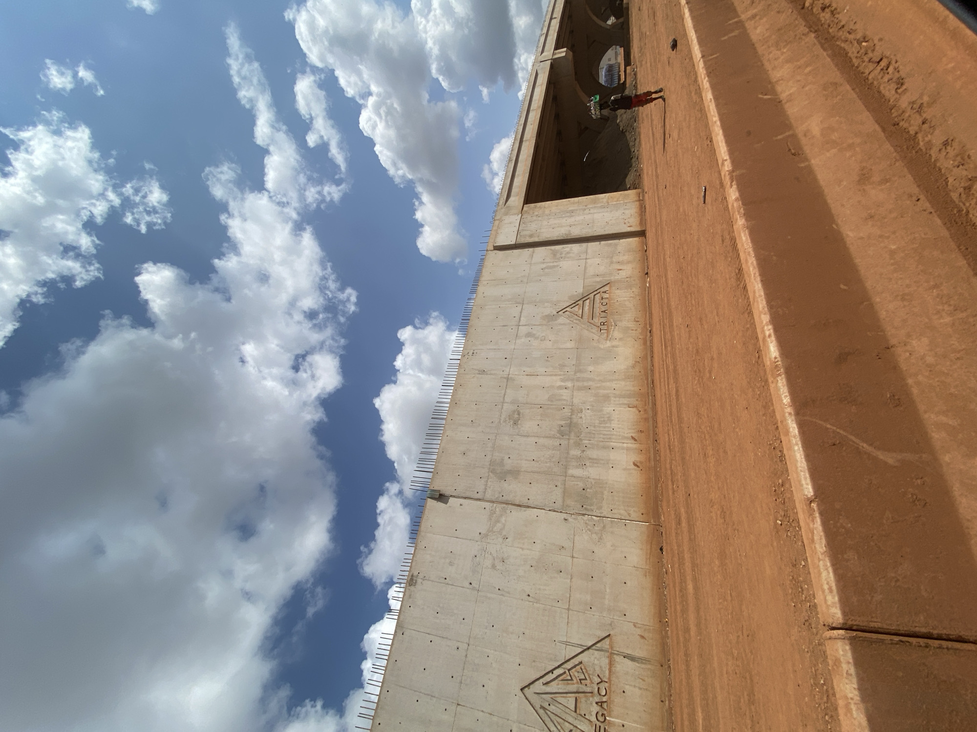 In Katambe, Nigeria, a construction site is depicted under a bright, partly cloudy sky. The foreground features an orange-brown dirt road with a concrete barrier. Dominating the scene is a large, unfinished concrete wall, part of a bridge or overpass, with exposed rebar along its top edge. Two triangular logos are embossed on the wall: "LEGACY" on the left and "TRIACTA" on the right. To the right of the wall, the underside of the bridge structure is visible, supported by curved concrete pillars. A lone individual, wearing red shorts and a dark shirt, walks along the dirt road, carrying a large bundle on their head. The environment is dusty and appears to be an active construction area.