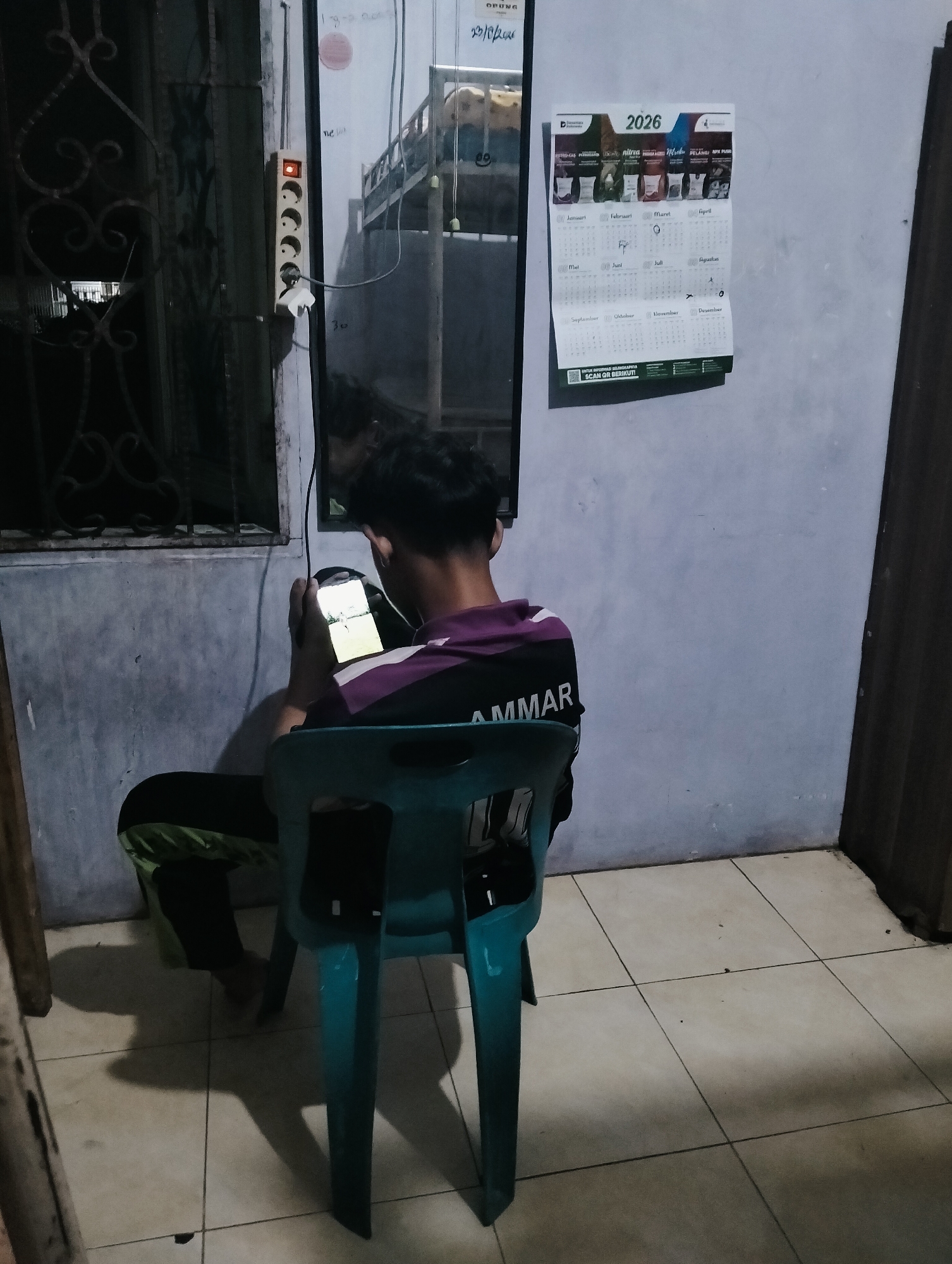 In a dimly lit room, a young male sits on a turquoise plastic chair, engrossed in a handheld gaming device. He is dressed in a dark shirt with purple and white accents, and green and black striped pants. The room appears to be a residential space in the City of Medan, Indonesia. The walls are a muted gray, and the floor is tiled with off-white squares. To the left of the frame, a metal security grate is visible over a window. A power strip with several outlets is mounted on the wall, with one outlet illuminated. A mirror reflects part of the room, showing bunk beds and a glimpse of another person. A calendar for the year 2026 is pinned to the wall on the right side of the frame. The overall atmosphere suggests a casual, indoor setting, possibly a dormitory or a shared living space.