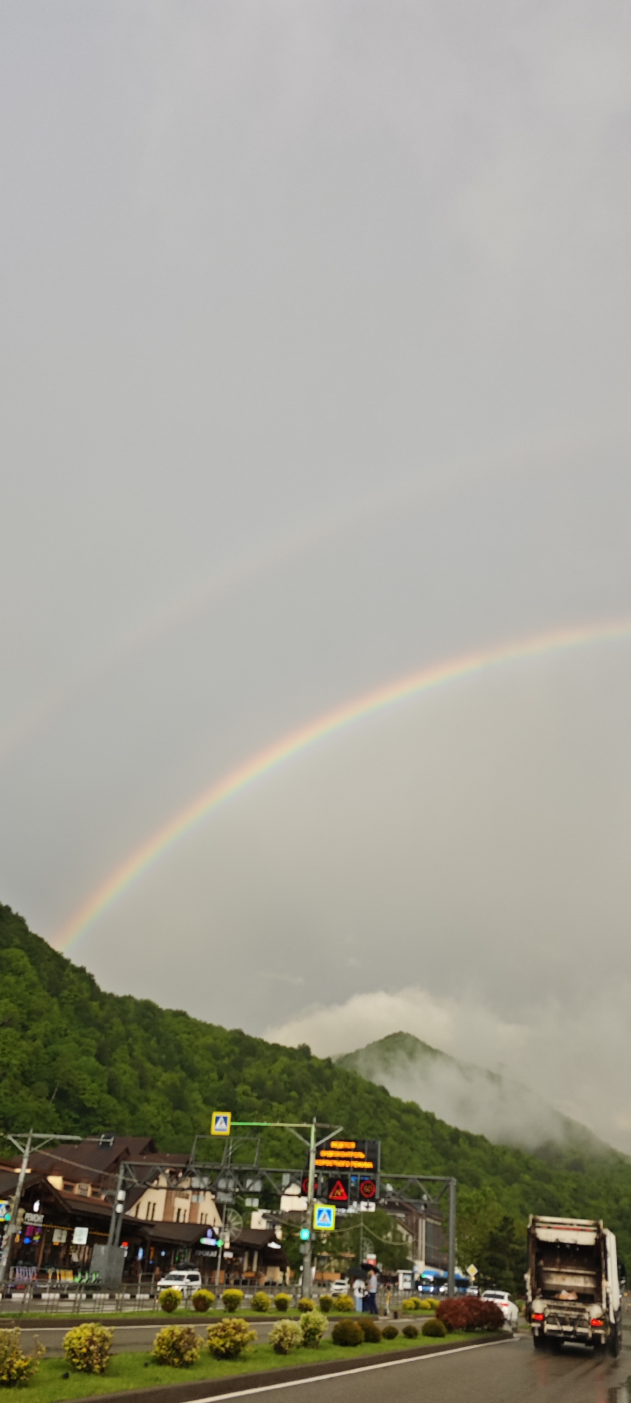 The image shows a landscape in Estosadok, Russia, with a prominent double rainbow arching across a cloudy sky above a green, tree-covered mountainside. In the foreground, there's a street with a row of buildings on one side, interspersed with pedestrian crossing signs and traffic signals. A garbage truck is visible on the right side of the road. People are walking along the sidewalk. The overall impression is a scene of a town nestled in a mountainous region, with a recent rainfall creating the rainbow effect.