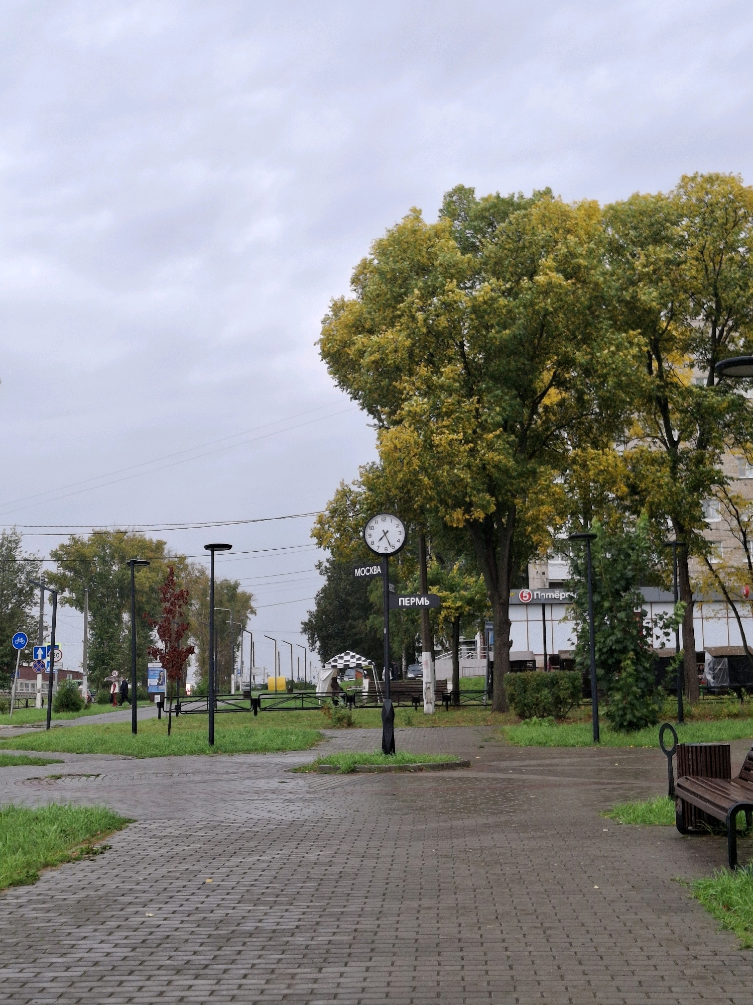 This image depicts a street scene in Chaikovsky, Russia, on an overcast day. A paved walkway, constructed with rectangular grey bricks, occupies the foreground and extends into the middle ground. The walkway is wet, suggesting recent rain, and is bordered by patches of green grass.
In the middle ground, a street lamp stands prominently to the left, and another is visible further down the street. A circular sign with a bicycle icon is attached to a pole near the street lamp, indicating a cycling path. Further along, there are traffic signs with numbers and arrows. Trees with yellow and green foliage are scattered throughout the scene, some lining the street and others in the background.
A large clock is mounted on a pole in the middle ground, displaying the time. Beneath the clock, there are two street signs, one reading "МОСКВА" (Moscow) and the other "ПЕРМЬ" (Perm). To the right of the clock, a building with multiple windows is visible, and a red circular logo with the number "5" and Cyrillic text "Пятёрочка" (Pyaterochka), a supermarket chain, is displayed on its facade.
Further into the background, more street lamps and trees are present. A checkered canopy structure and a slide are visible near the right edge of the frame, suggesting a children's play area or recreational space. A wooden bench with dark metal legs is situated on the right side of the walkway in the foreground. A few individuals are walking in the middle ground, and parked vehicles can be seen in the distance. The overall environment appears to be a residential or public area within a city.