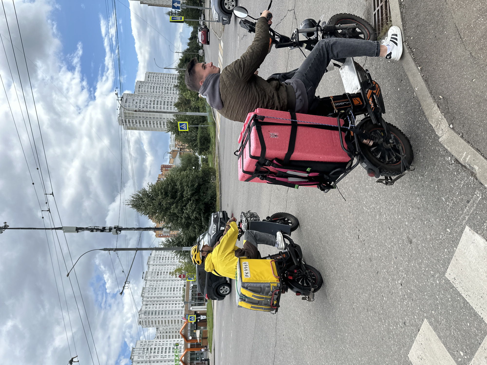 Two delivery drivers on electric scooters are riding on a city street in Podolsk, Russia. The driver in the foreground, on the right, is a young man wearing a dark green jacket and a grey hoodie, with a large pink insulated delivery box attached to the back of his scooter. The driver in the background, on the left, is wearing a bright yellow jacket and a yellow helmet, with a yellow delivery box on his scooter. Both scooters have cargo compartments. The street has asphalt pavement and is marked with a pedestrian crossing. In the background, there are multi-story apartment buildings, trees, and utility poles with overhead wires. The sky is cloudy.
