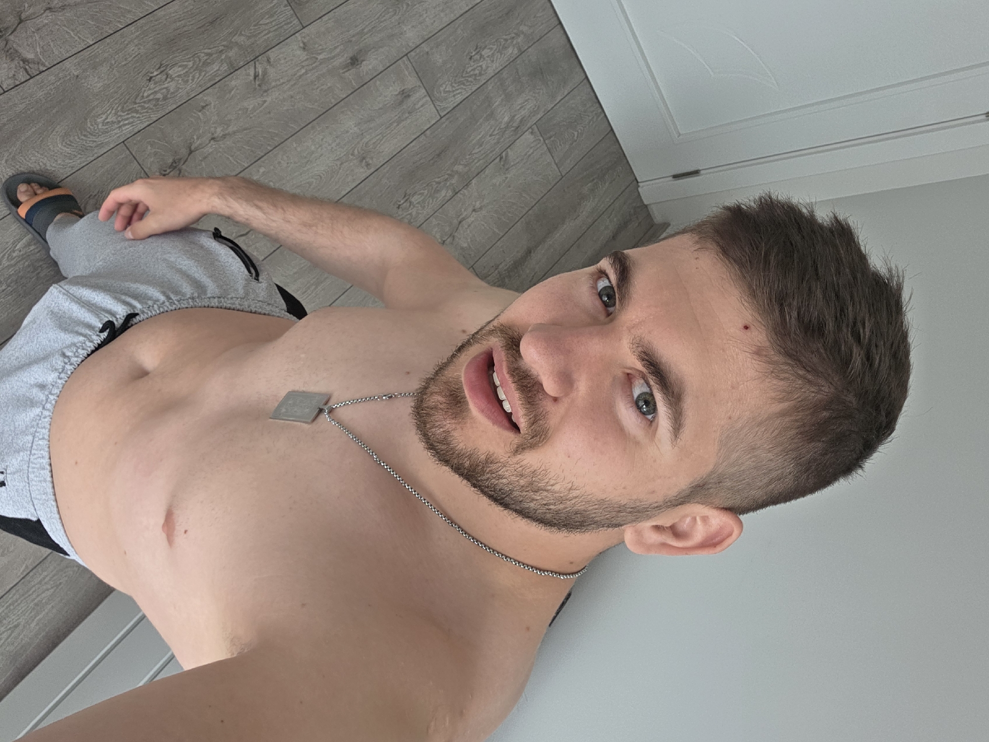 A medium shot features a fair-skinned male, shirtless and angled towards the camera, with grey sweatpants and a sandal visible in the lower left. He wears a silver chain necklace with a square pendant. His beard is short and trimmed, and his hair is dark and neatly cropped. A small red mark is visible on his forehead. The background consists of grey wood-look flooring and a plain, light-colored wall with a white door in the upper right. The setting is indoors. The image was not taken in Yagodovo, Bulgaria because the setting doesn't match Yagodovo's known indoor environments.