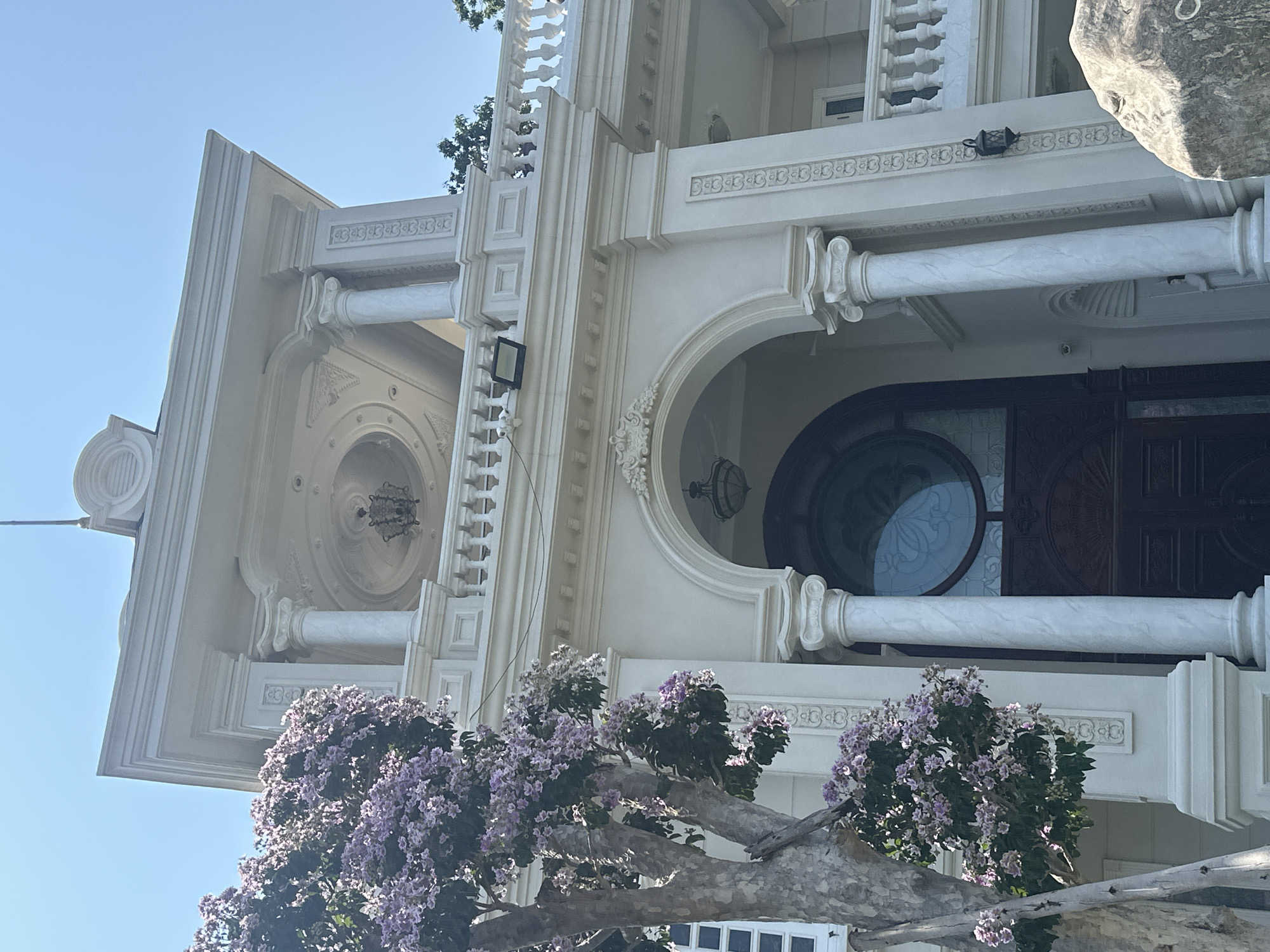 A low-angle shot reveals the ornate facade of a building in Nam Nha Trang Ward, Vietnam. The building's architectural style is classical, featuring off-white stucco, columns, balustrades, and arched doorways. The primary entrance is a dark wood door with a circular glass window. A flowering tree with purple blossoms partially obscures the lower portion of the building.