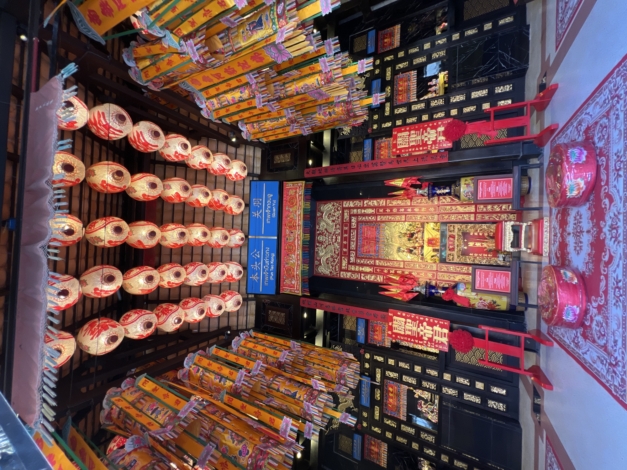 This is a low-angle, eye-level shot of the interior of a temple in Baan Hua Thanon, Thailand, likely dedicated to Chinese deities. The scene is elaborately decorated with red and gold accents, traditional Chinese lanterns, and banners.
In the foreground, a red patterned carpet covers the floor, upon which two cylindrical red ottomans with floral designs sit. To the left, a red wooden stand with a large red flower adorns it. A dark wooden cabinet with intricate gold detailing lines the walls.
Dominating the central background is an altar area, adorned with gold carvings and religious iconography. Two golden statues of deities are visible on the altar. Red fabric banners with Chinese characters hang from the ceiling, as do numerous round, white and red lanterns. Signs with blue and white lettering, bearing Thai and Chinese text, are also present.
The overall impression is one of rich cultural and religious significance, with a strong emphasis on traditional Chinese aesthetics. The lighting suggests it might be daytime, with artificial lights from the lanterns illuminating the space. There are no people visible in the image.