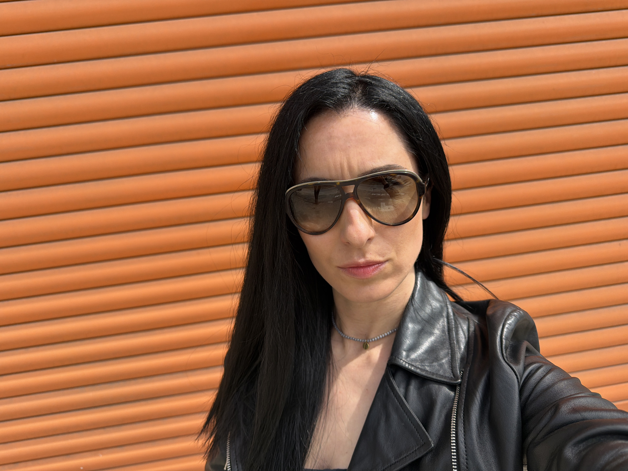 A woman with long black hair stands in front of an orange horizontally ribbed wall, wearing a black leather jacket, a gray beaded necklace with a gold pendant, and large aviator sunglasses. The location is unknown; there is no indication it is in the city of Pleven, Bulgaria.