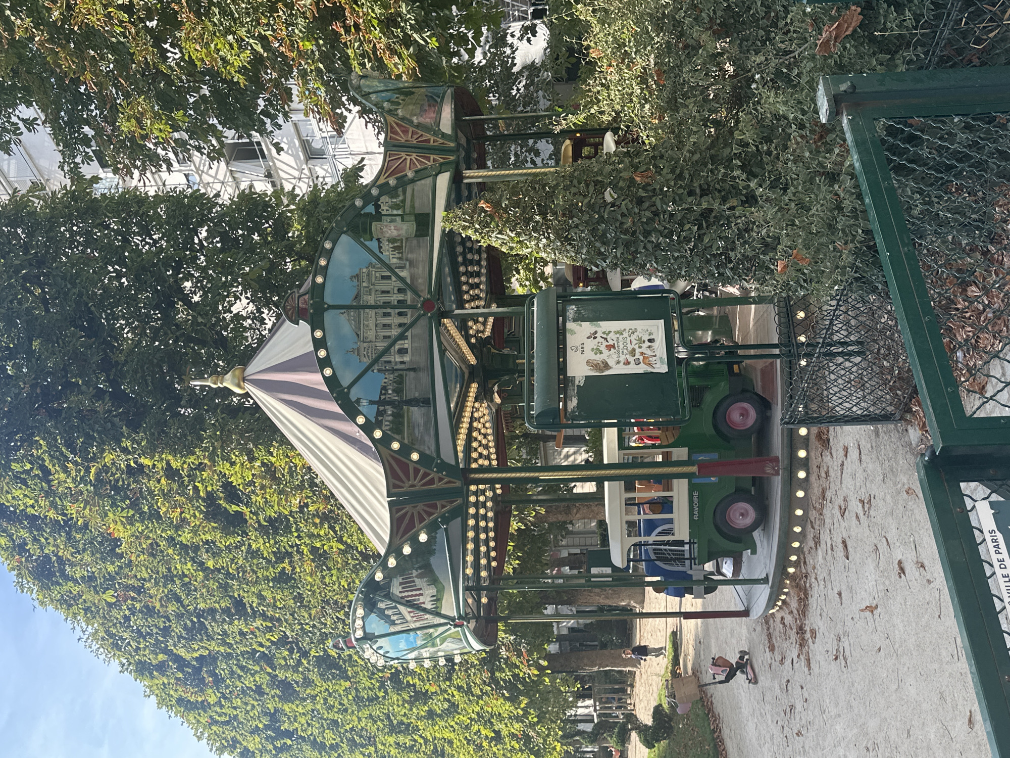 The image depicts an outdoor scene in Paris, France, likely a park or public square, characterized by lush green trees and a paved pathway. Dominating the frame is a colorful, carousel-like amusement ride, painted in green and adorned with numerous small, bright lights. The ride features decorative elements and painted scenes on its facade.
In the foreground, a child is visible, seemingly playing or interacting with something on the ground. Another person, possibly an adult, is also present, standing further down the pathway. The presence of a fence with a sign that reads "LA VILLE DE PARIS" (The City of Paris) strongly indicates the location. The sunlight suggests it is daytime, and the dappled lighting through the trees hints at a pleasant, possibly sunny, day. The overall atmosphere appears relaxed and pleasant, suggesting a leisurely outing in a public space. The ride itself, with its classic design and lighting, evokes a sense of nostalgia and family-friendly fun.