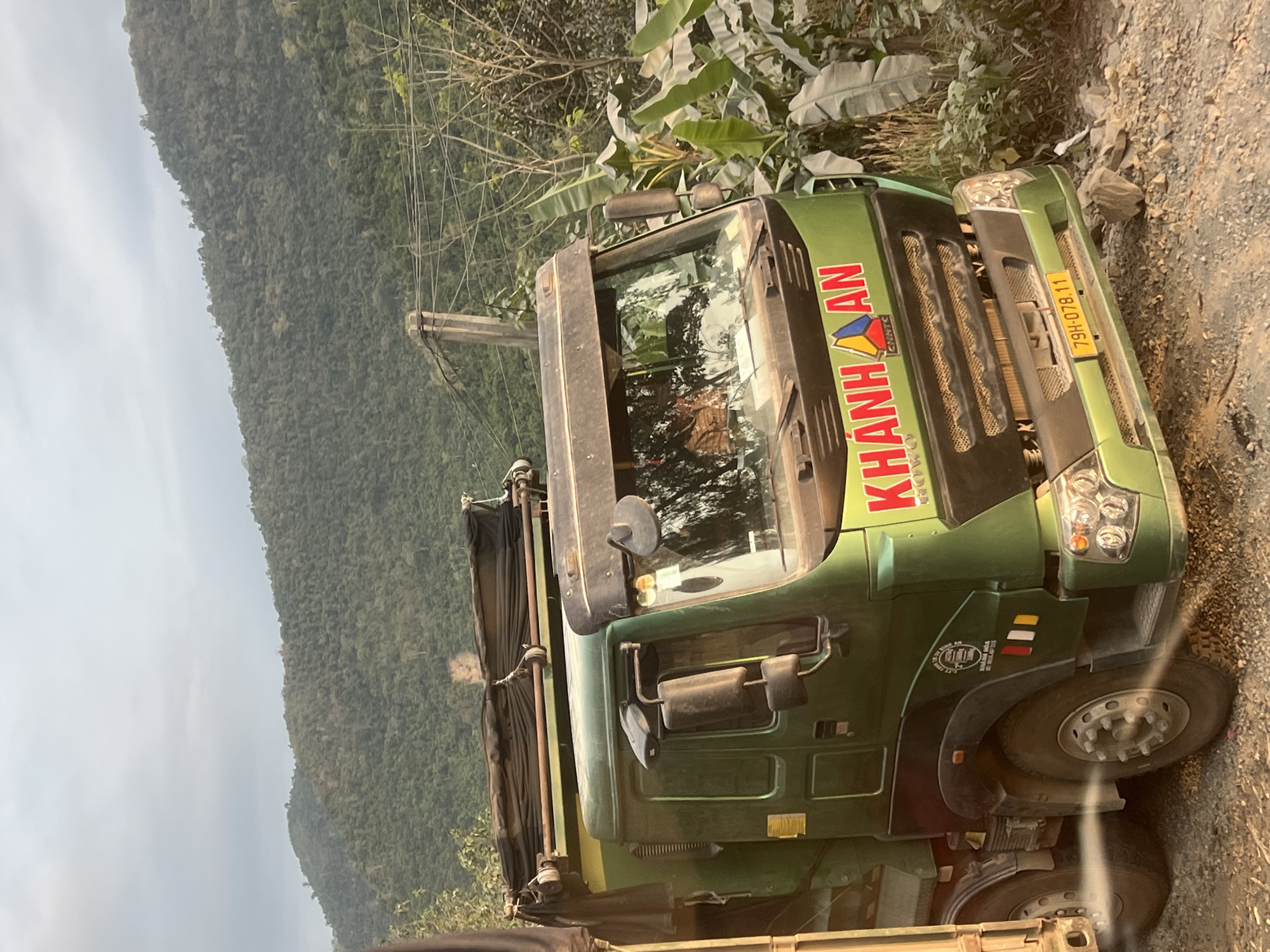 A green "KHANH AN" truck with license plate 79H-078.11 is parked on a dirt or gravel road. The truck's front end is visible, showing the grill, headlights, and front tires. The road is adjacent to a lush green mountainside and foliage. The photo was taken in Aê Thi, Vietnam. A telephone pole with wires is also in the background.